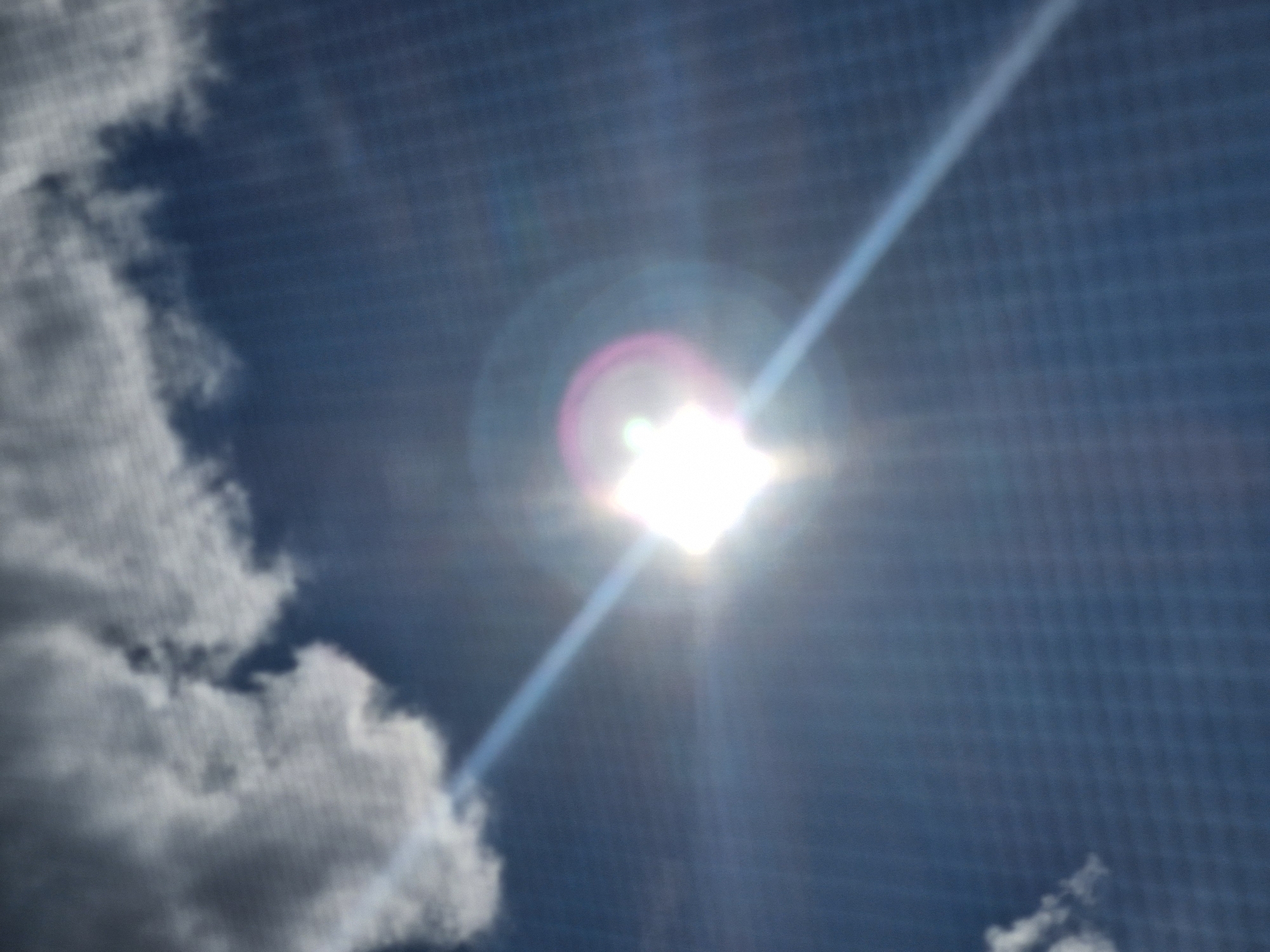 The image depicts a sunburst effect in a blue sky with clouds. The sun is positioned slightly off-center, casting a bright glare and a light beam across the sky. Some clouds are visible, mostly in the upper left portion of the image and a few smaller ones scattered in the lower left. The background has a faint grid pattern, suggesting the image was taken through a screen or window. There are no identifiable landmarks or elements that would place this scene specifically within the city of Kazan, Russia. The scene is devoid of people or any other recognizable objects.