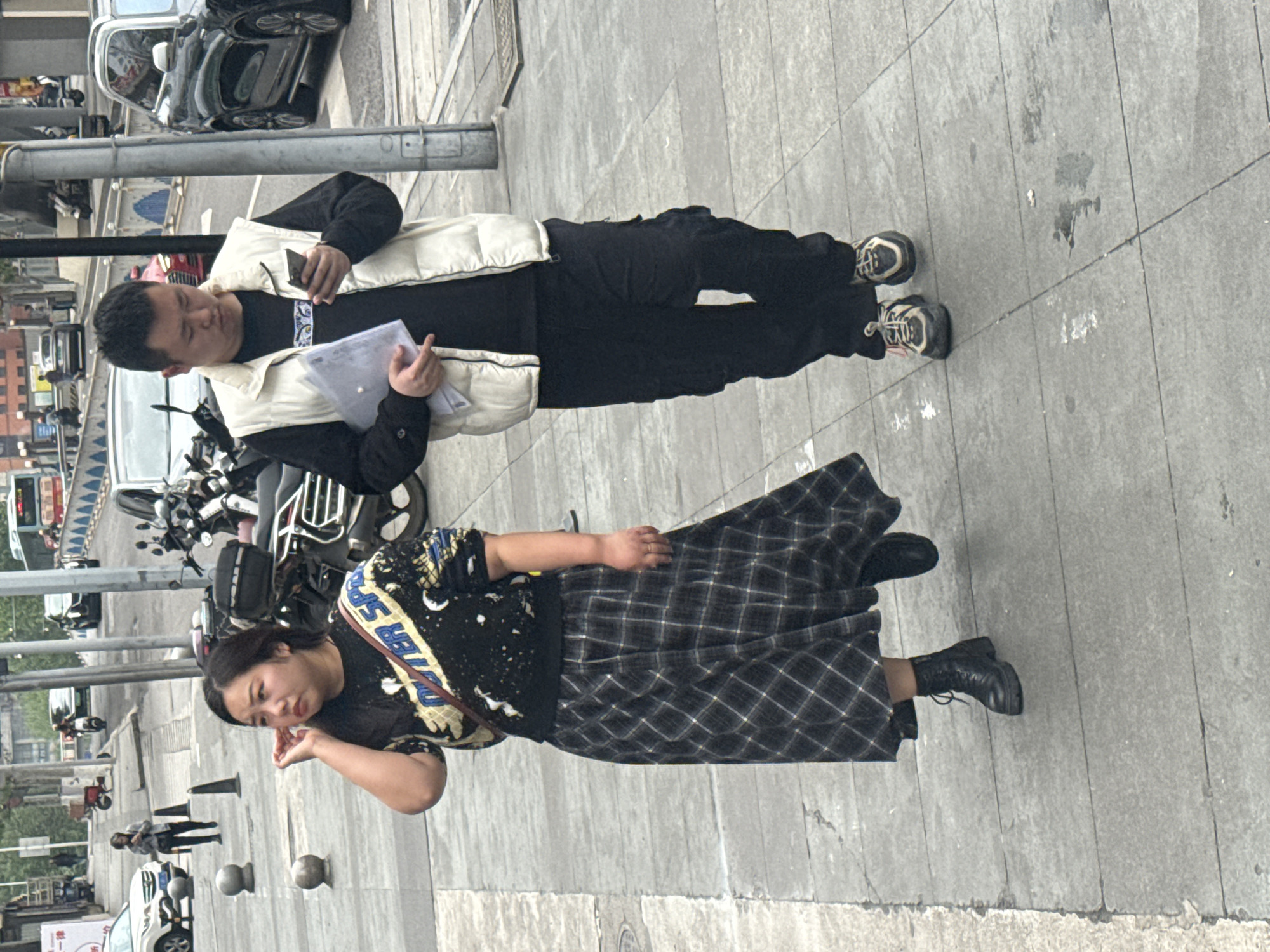 On a street in Yuzhong District, China, a man wearing a black long-sleeve shirt, black pants, a beige puffer vest, and sneakers, holds documents and a phone while standing next to a motorcycle. Nearby, a woman in a black patterned t-shirt, a long plaid skirt, and black boots touches her face while looking down the street. In the background are parked cars, buildings, and people walking on the sidewalk. The ground is paved with grey stone slabs.