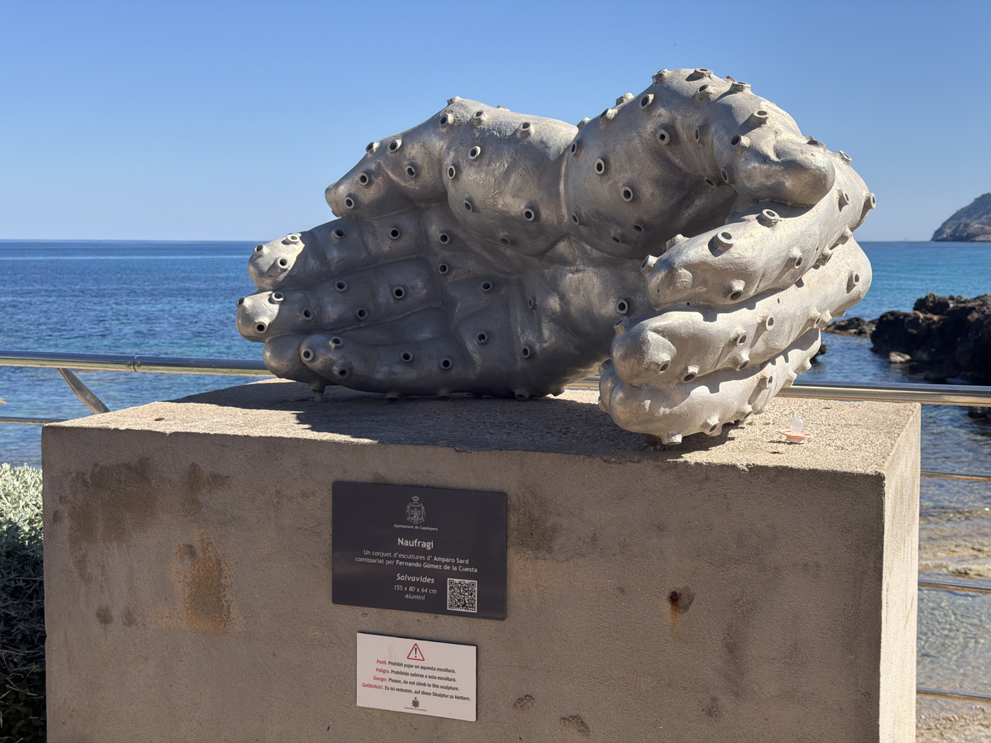 A metal sculpture resembling a mass of life preservers is on display in Capdepera, Spain. The sculpture, titled "Naufragi" (Shipwreck), is placed on a concrete plinth near the seaside, as indicated by the view of the ocean and coastline in the background. A plaque with information about the artwork and artist is attached to the plinth. A warning sign advising people not to climb on the sculpture is also visible.