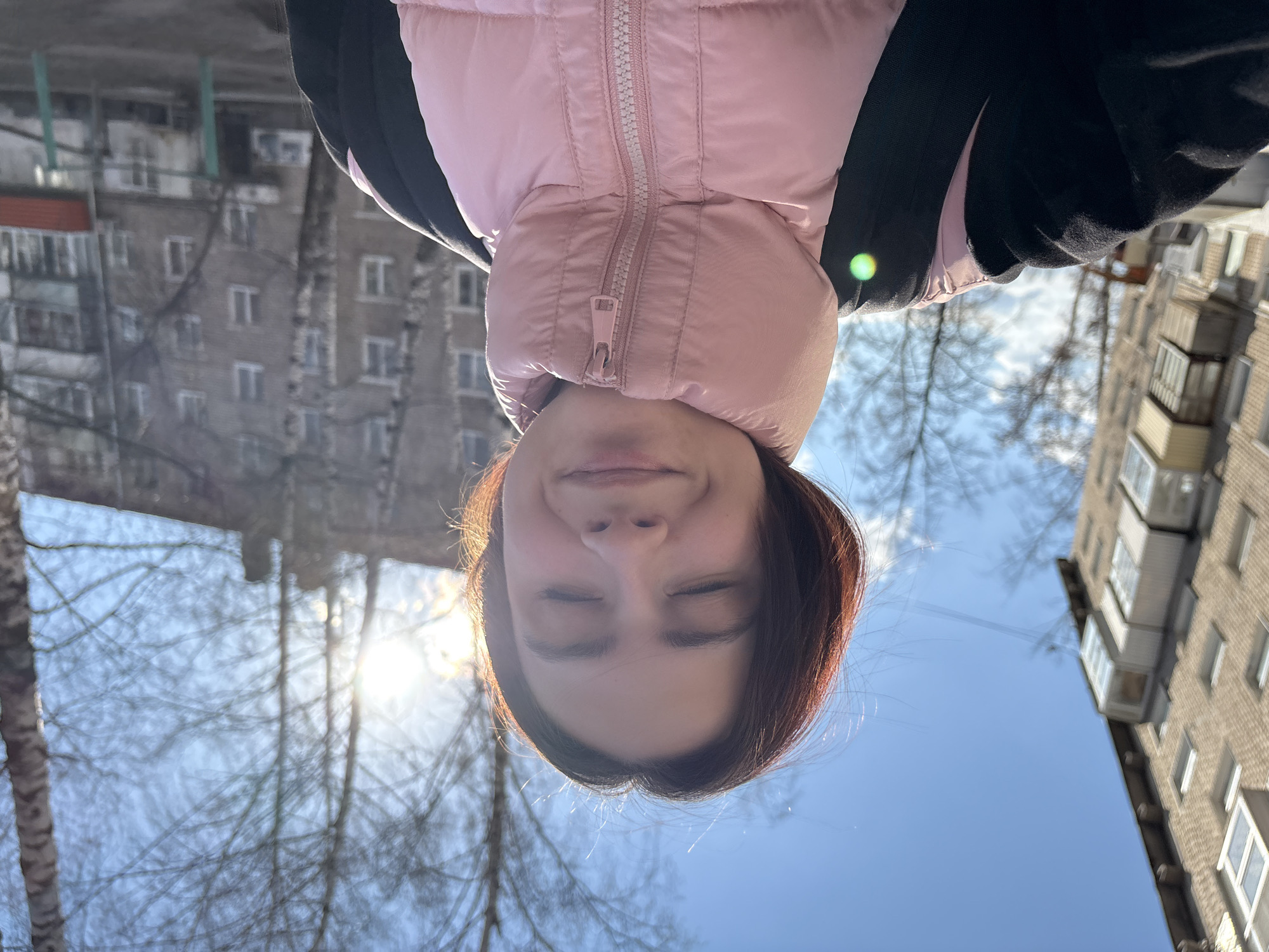 This rotated image (90 degrees clockwise) captures an outdoor, low-angle selfie, depicting a person looking upwards towards a clear, bright blue sky. The central subject is seen upside down, wearing a light pink puffer jacket with a prominent zipper. They have dark brown hair, and their eyes are closed with a gentle smile on their face, conveying a sense of peace or enjoyment of the sunshine.
The background shows an urban or suburban setting, consistent with Chaikovsky, Russia. On both sides of the subject, multi-story brick residential buildings, likely apartment blocks, are visible, some featuring balconies. Bare, leafless trees are scattered throughout the scene, suggesting a winter or early spring setting. Bright sunlight creates a prominent lens flare in the upper-left portion of the frame (when viewed right-side up), indicating a sunny daytime. There is no visible text in the image.