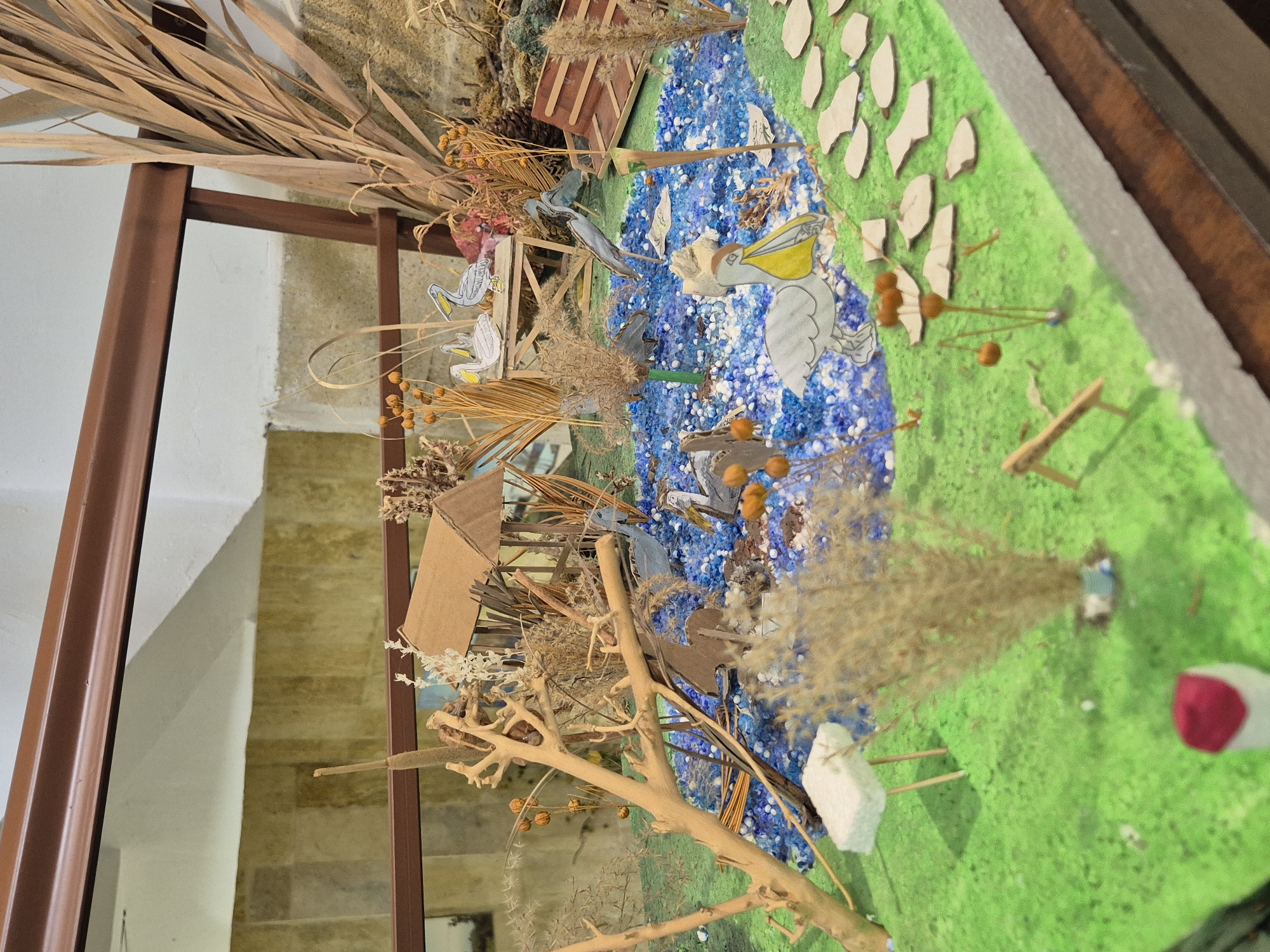 This image depicts a diorama representing a wetland ecosystem. Key elements include a body of water simulated with blue materials, green "grass" terrain dotted with model trees and vegetation. Model birds, specifically pelicans and other waterbirds, are placed in and around the water. A small, constructed structure (possibly a hut or bridge) is also present. Dried plants and branches are used for additional landscaping effects.
While not explicitly stated in the image, given the prompt, the diorama is most likely intended to represent the Srebarna Nature Reserve in Bulgaria, a UNESCO World Heritage Site known for its diverse birdlife.