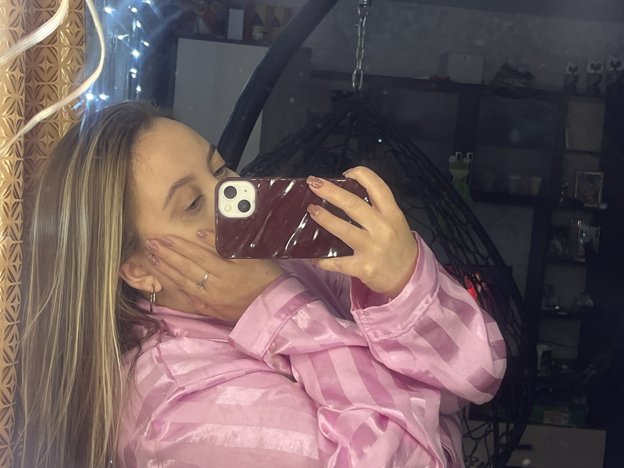 A woman in a pink striped pajama top is taking a selfie in a mirror. Her face is partially obscured by her phone and hand. The background includes fairy lights, a patterned curtain, a hanging chair, and a dark shelving unit with various objects on it. The shelving unit and overall scene suggest an indoor setting, likely a home. There is no indication of the city of Novopokrovskaya, Russia.