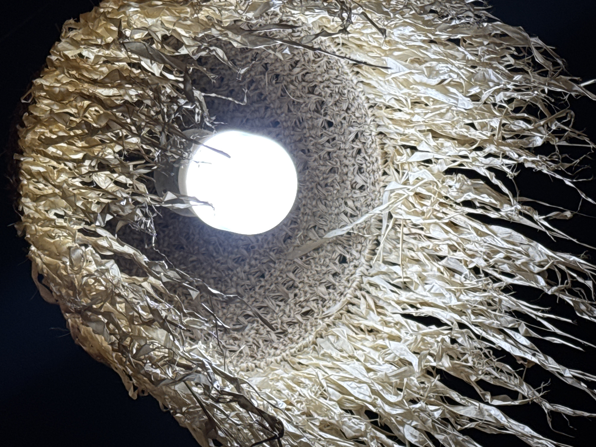 This media file is a close-up, low-angle shot of a ceiling lamp featuring a unique, rustic lampshade. The lampshade is constructed from what appears to be dried, light-colored plant material, possibly straw or reeds, which are woven and twisted into a textured, conical shape. Long, thin strips of the same material hang loosely from the outer edges, creating a fringed effect that sways outwards. The material is somewhat translucent, allowing the light from the bulb within to create a warm, diffused glow.
The central subject is the illuminated lightbulb, a bright white, spherical object positioned at the apex of the lampshade. The texture of the lampshade is intricate, with many small, interwoven pieces forming a dense mesh around the light source, transitioning to the more flowing, ribbon-like strands on the exterior. The background is a dark, solid black, emphasizing the light and the texture of the lampshade. The overall mood is natural and organic, with a touch of handcrafted artistry. The lighting suggests it might be evening or indoors, as the darkness of the background is absolute. There is no visible text. The location context provided is Kuta, Indonesia.