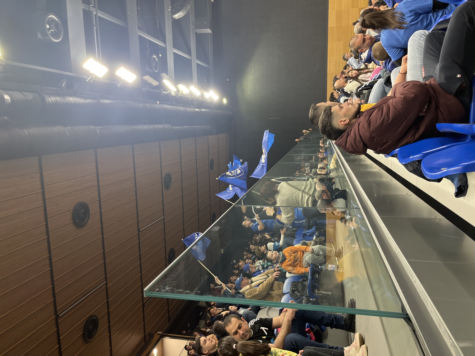 The image shows an indoor scene with spectators seated in an arena-like setting. There are rows of blue seats filled with people, many wearing blue shirts and holding blue flags. The arena has a glass barrier separating the seating area from the background. The background features a wall with wood paneling, black circular vents, and a black upper section with lighting fixtures. The scene is likely a sporting event or a similar public gathering. The location is likely in Sofia, Bulgaria, based on the flag design being that of PFC Levski Sofia.