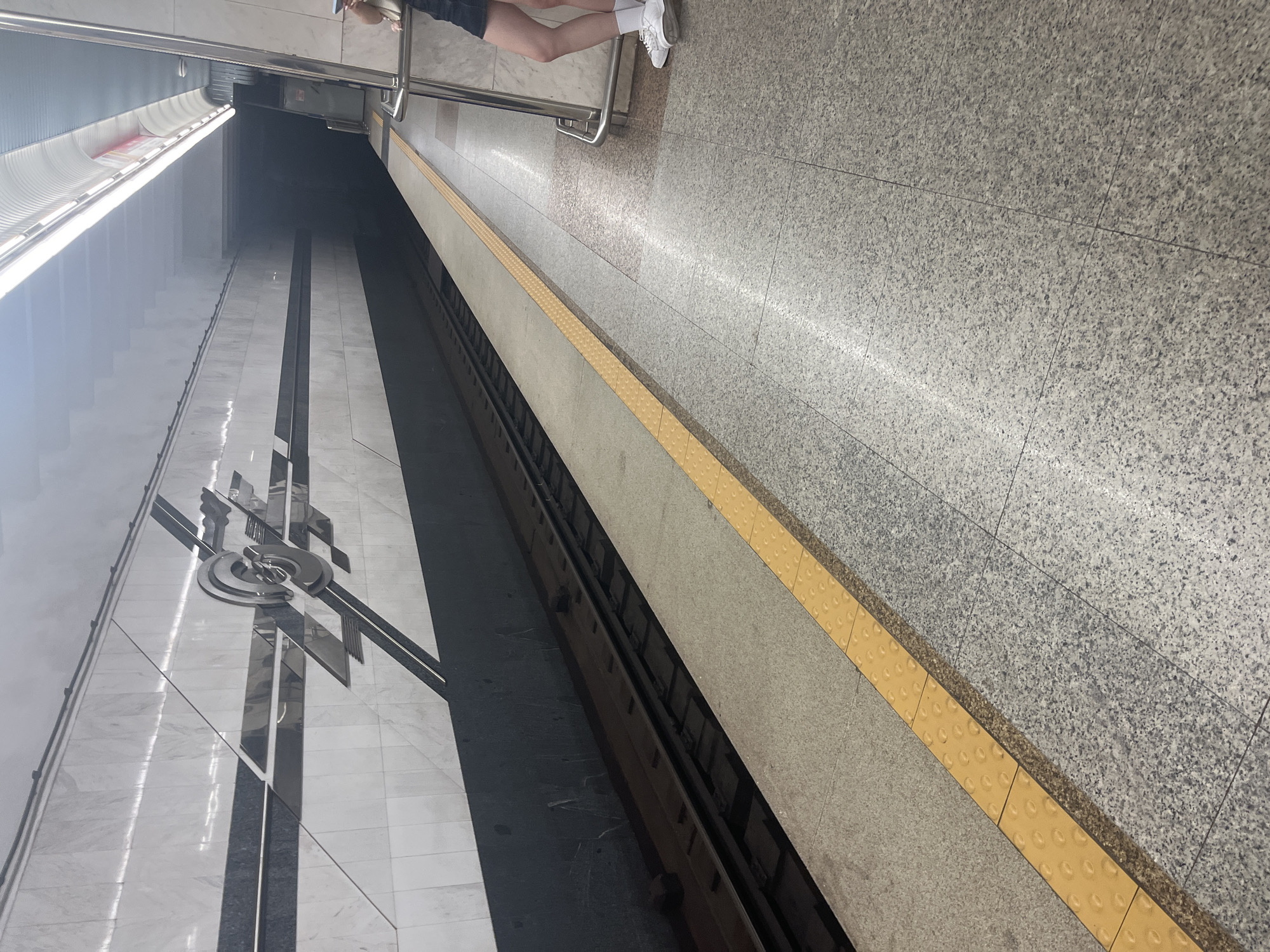 The image depicts a subway station in Minsk, Belarus. The scene shows a subway platform with a yellow tactile paving strip along the edge, separating it from the train tracks. The platform floor is made of granite tiles. The walls of the station are clad in white marble with dark grey decorative accents and a metallic emblem. Further down the platform, the tunnel entrance is visible, shrouded in darkness. A person is standing on the platform to the right, leaning against a railing, wearing white sneakers, white socks, and a dark skirt. The lighting suggests it is indoors, typical of a subway environment.