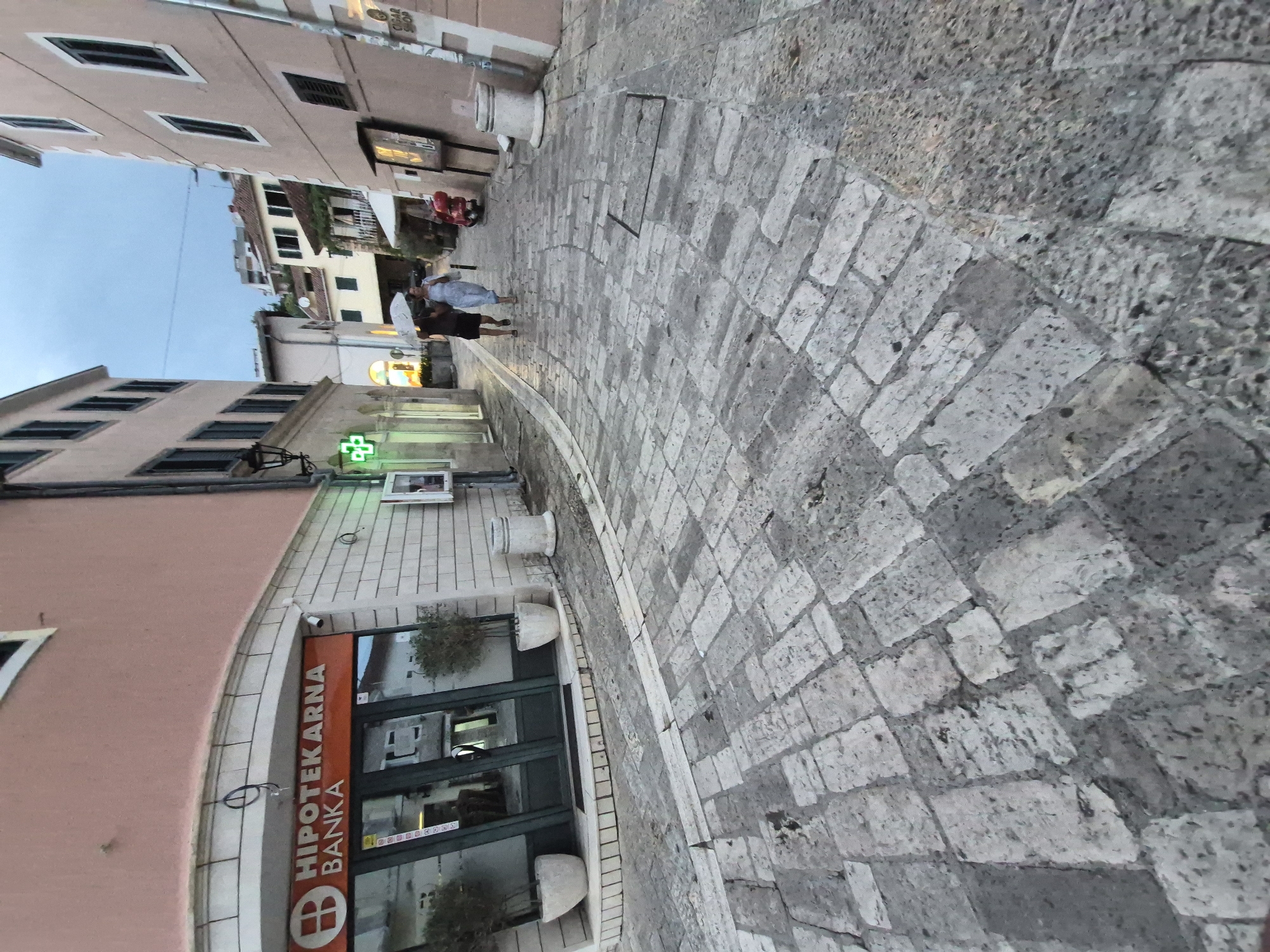 In Herceg Novi, Montenegro, a narrow, cobblestone street curves between two buildings. On the left, a salmon-pink building houses a "HIPOTEKARNA BANKA" branch with a large glass entrance, flanked by two potted trees. A CCTV camera is mounted above the entrance. The building to the right is a lighter beige color, with several windows and a sign that reads "CIGAR SHOP".
Two individuals are walking away from the camera down the street. The person in front is wearing a light-colored dress and holding a white umbrella. The person behind them is wearing dark shorts and a dark shirt. A red scooter is parked near the building on the right, further down the street. The sky is overcast, suggesting it may be evening or raining. The street is illuminated by a green cross sign, possibly indicating a pharmacy, and other ambient lighting from shops. The cobblestones appear worn and uneven, characteristic of historic European streets.