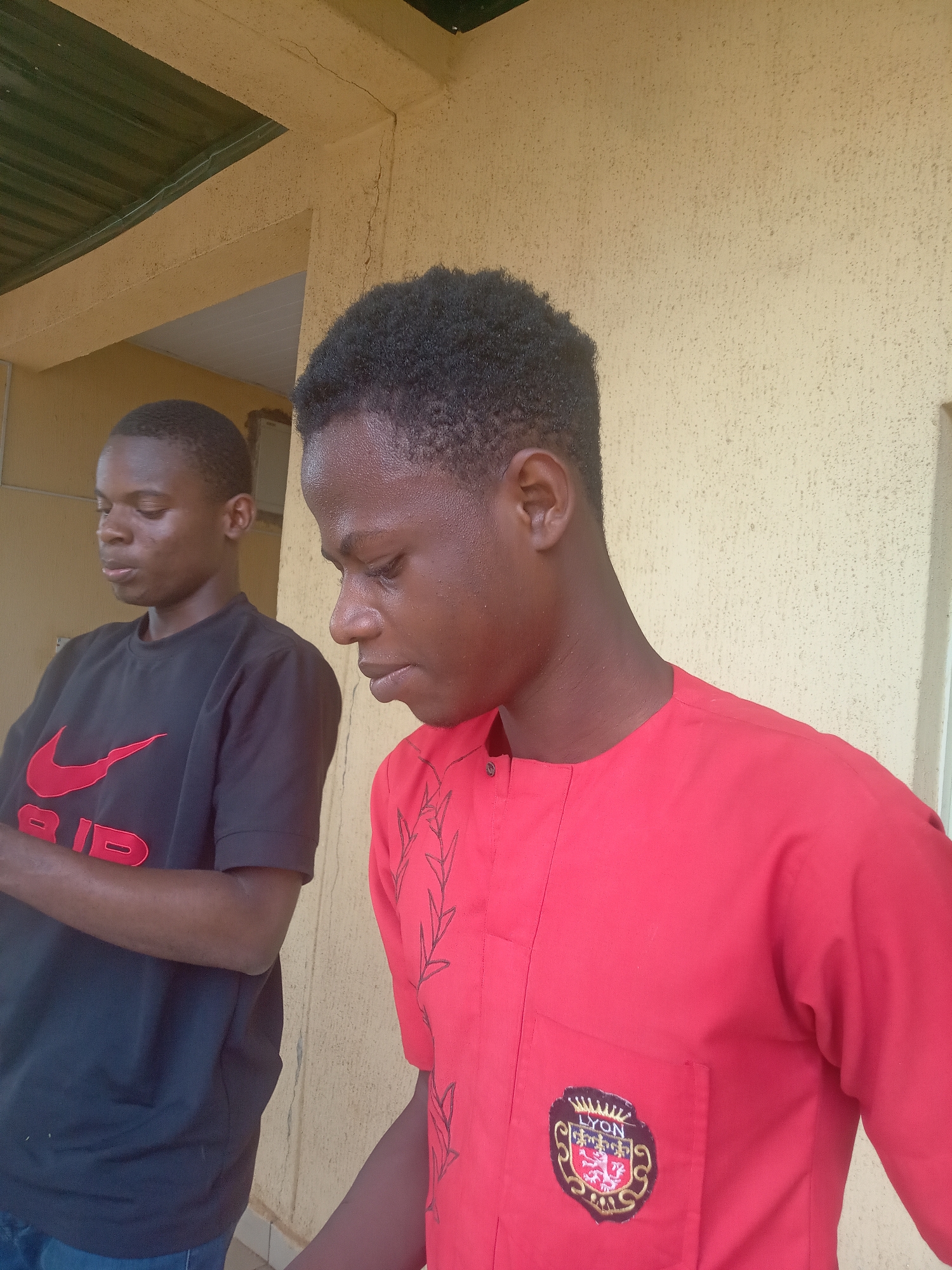 Two young men of African descent are captured in a daytime, outdoor setting in Nkwelle, Nigeria. The image is taken from a slightly low angle, focusing on the two individuals.
The man in the foreground is positioned slightly to the right, facing downwards and to his left, revealing his profile. He has short, dark, curly hair and is wearing a vibrant red traditional shirt with a band collar and three small buttons at the neck. The shirt features intricate black embroidery of leafy branches on the front, extending from the shoulder downwards. On the left chest pocket, there is a prominent crest. The crest is a shield design with red and gold elements, featuring a crowned lion rampant and other symbols. The word "LYON" is clearly visible across the top of the crest. His skin appears smooth and well-lit.
Behind him, to the left, stands another young man, partially obscured and slightly out of focus. He is also looking downwards or away, with a neutral expression. He wears a black t-shirt with a large, bright red Nike "swoosh" logo prominently displayed on the chest. Below the swoosh, the letters "AIR" are visible in red. His arms are crossed in front of him.
The background consists of a textured, light yellowish-beige wall, likely plaster or stucco, showing some hairline cracks, particularly in the upper right. Above the wall, a dark green corrugated roof or awning is visible, suggesting a covered outdoor area. The lighting is bright and natural, indicating daylight, with soft shadows. The overall mood is calm and observational.