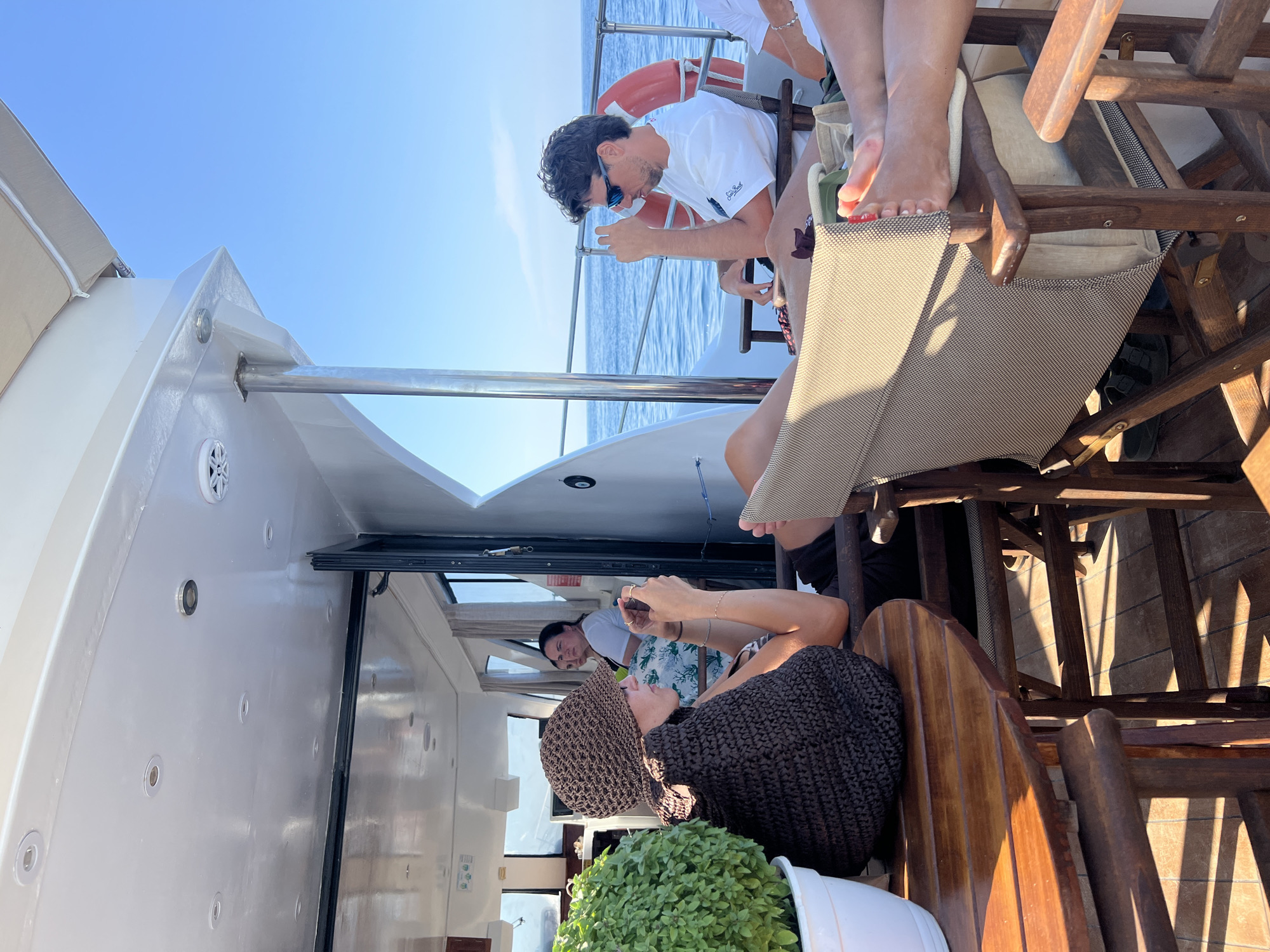On a sunny day, a group of people relax on the deck of a boat on the Aegean Sea, near the coast of Skopelos, Greece. The boat appears to be a small yacht or ferry with a white superstructure and a wooden deck. In the foreground, a woman wearing a brown crocheted hat and a dark knitted garment is seated at a wooden table, holding a smartphone in her hands. To her right, a man in a white t-shirt and sunglasses is seated, looking out at the sea. A life preserver is visible on the railing behind him. Other passengers are seated in chairs, some with their legs propped up. The water is calm and blue, and the sky is clear. Green foliage is visible in a white pot on the left side of the image.