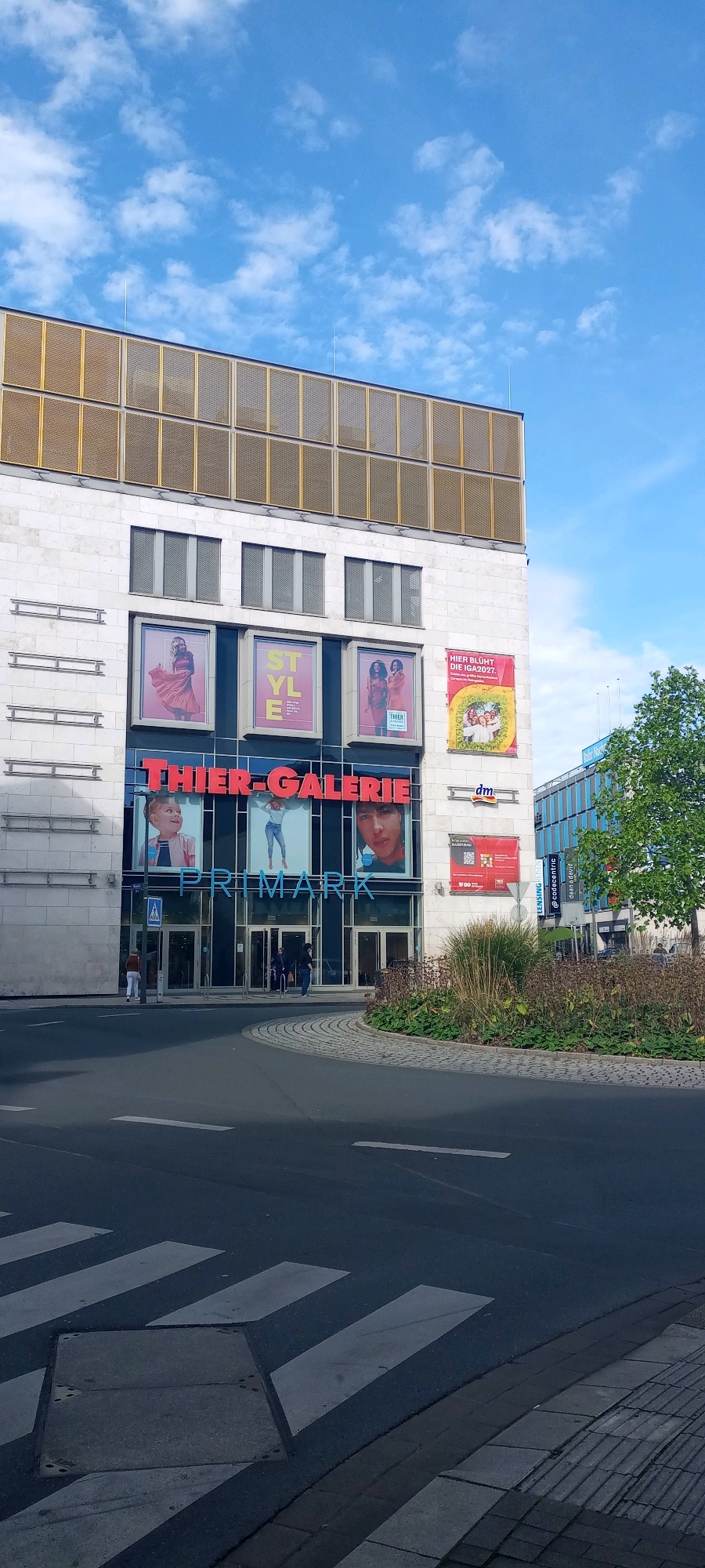 The image depicts the exterior of a multi-story commercial building, identified by a large red sign as "THIER-GALERIE". Below this sign, another prominent blue sign reads "PRIMARK", indicating a clothing store. The building features large glass windows and several advertising panels displaying fashion-related imagery. To the right of the Primark entrance, there is a smaller sign for "dm", a drugstore. The building is situated in a city environment with a street in the foreground featuring a crosswalk. A tree and other greenery are visible to the right of the building. The sky is blue with scattered clouds. The building appears to be located in Dortmund, Germany, as suggested by the context of similar images.