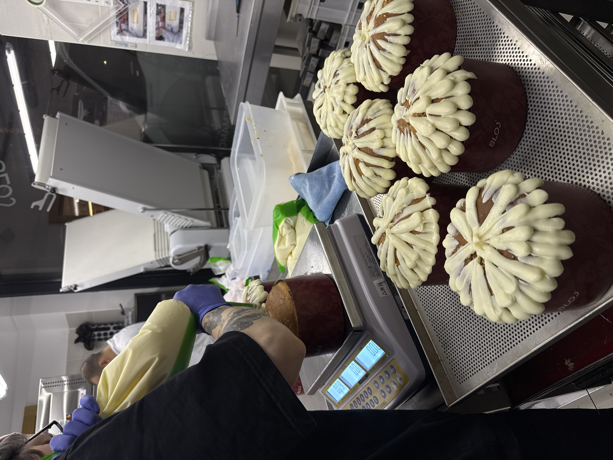 A person wearing blue gloves is weighing a decorated cake on a digital scale. The scale reads 0.00. Another person in a white shirt is visible in the background. A tray of similar cakes decorated with piped icing is positioned to the right of the scale. The cakes are labeled "SLOJ." The scene appears to be taking place in a commercial kitchen. The location is Belgrade, Serbia.