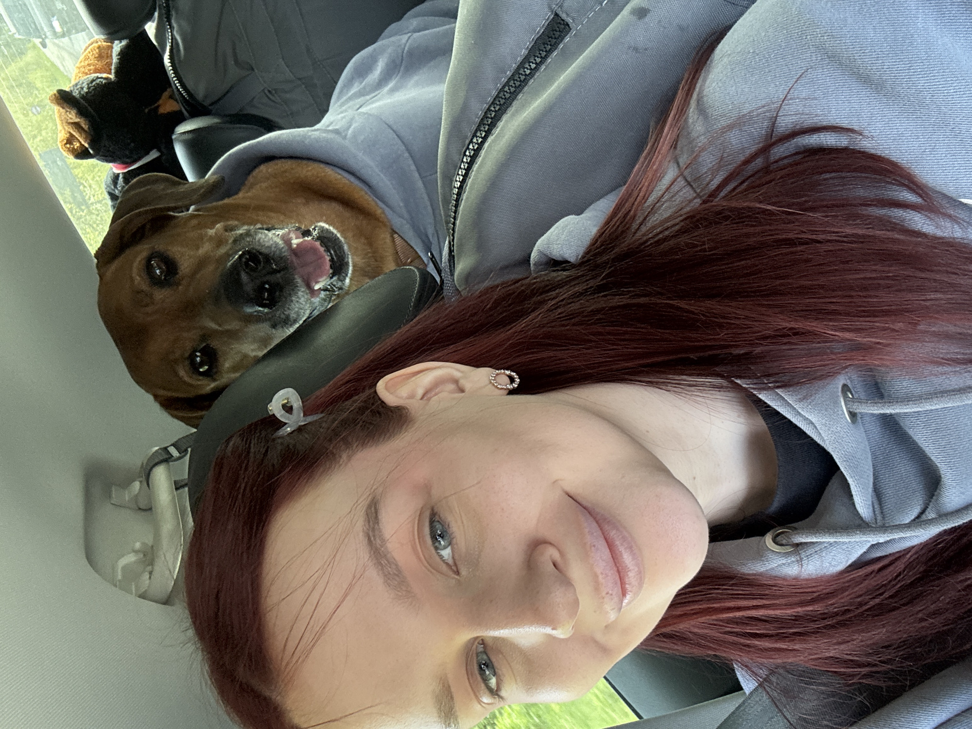 A selfie photograph shows a woman and a dog in the interior of a vehicle. The woman has light skin, red hair, and is wearing an earring. A clear hair clip is visible in her hair. She is looking at the camera and smiling.
The dog is medium-sized with brown fur. It has its mouth open, appearing to smile. The dog is wearing a gray hoodie.
The scene appears to be inside a car. The background shows parts of the car's interior, including a headrest, window, and seat. The location in the city of Balakhna, Russia cannot be determined from the visual information in the photograph.