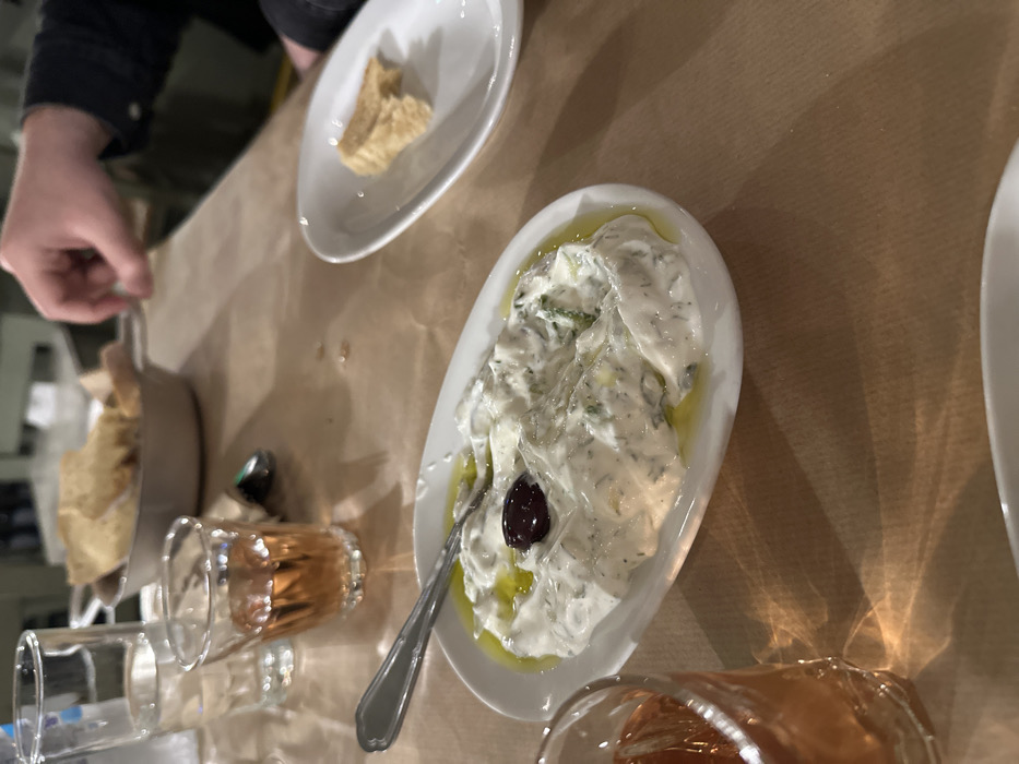 This media file captures a close-up, slightly overhead view of a table set for a meal, likely in a restaurant or taverna in Nea Erithrea, Greece. The scene is dominated by food and drinks, suggesting a relaxed dining experience.
In the foreground and central to the image is an oval white dish filled with a creamy, pale green dip, typical of tzatziki, garnished with a single dark olive and drizzled with olive oil. A metal spoon rests within the dip. To the left and slightly behind this dish, another similar white oval dish holds pieces of golden-brown bread or pita. Further to the left, a basket filled with more bread is visible, with a person's hand reaching towards it, implying they are about to take a piece or are placing something down.
Several glasses are scattered on the light brown paper-covered table. Two glasses contain a rosy or amber-colored liquid, possibly rosé wine or another beverage. Another empty clear glass is also visible. The lighting appears warm and artificial, characteristic of an indoor dining setting, with reflections visible on the table surface, likely from overhead lights. The casual paper tablecloth, the style of the dishes, and the presence of tzatziki and pita strongly indicate a Greek culinary context, aligning with the specified location of Nea Erithrea, Greece. No visible text is present in the image.