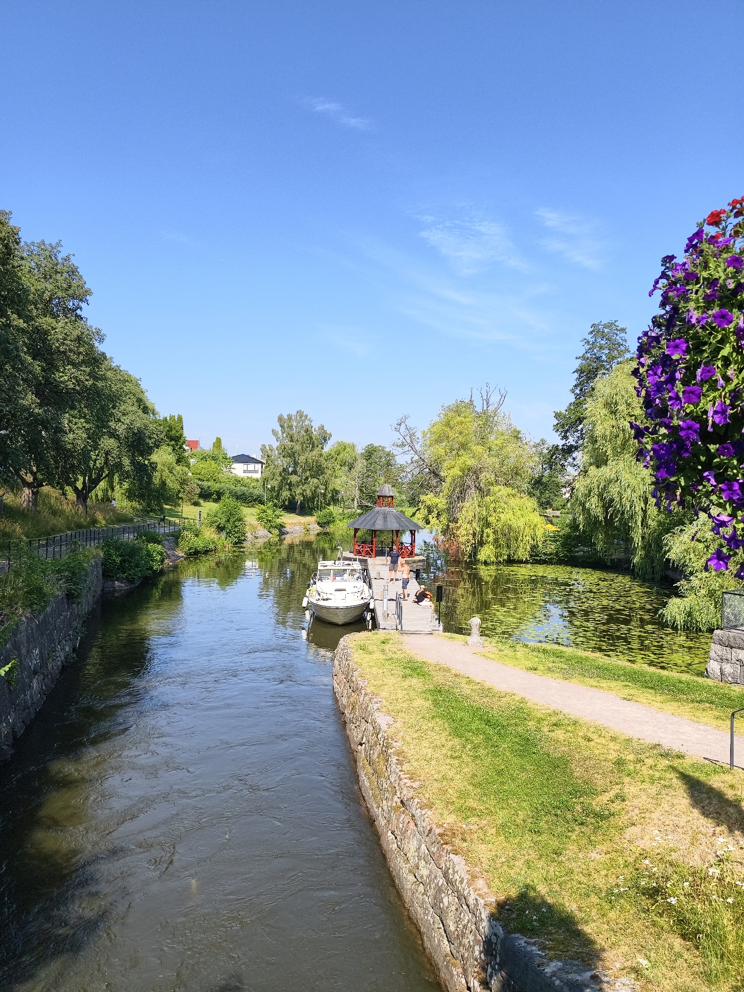 The image captures a scenic view of the Göta Canal in Linköping, Sweden. The canal, with its stone-lined banks, flows through a verdant landscape. On the right bank, a pathway runs alongside the water, bordered by lush green grass and punctuated by blooming purple flowers cascading from an overhead structure. A small white boat is docked at a wooden pier where several people are visible. Further into the scene, the canal opens up into a wider section covered with lily pads, and beyond that, a picturesque gazebo with a red roof stands near the water's edge. The left bank of the canal features a walking path shaded by mature trees, with houses visible on the elevated ground in the distance. The sky above is a clear, bright blue with a few wispy clouds, indicating a pleasant day.