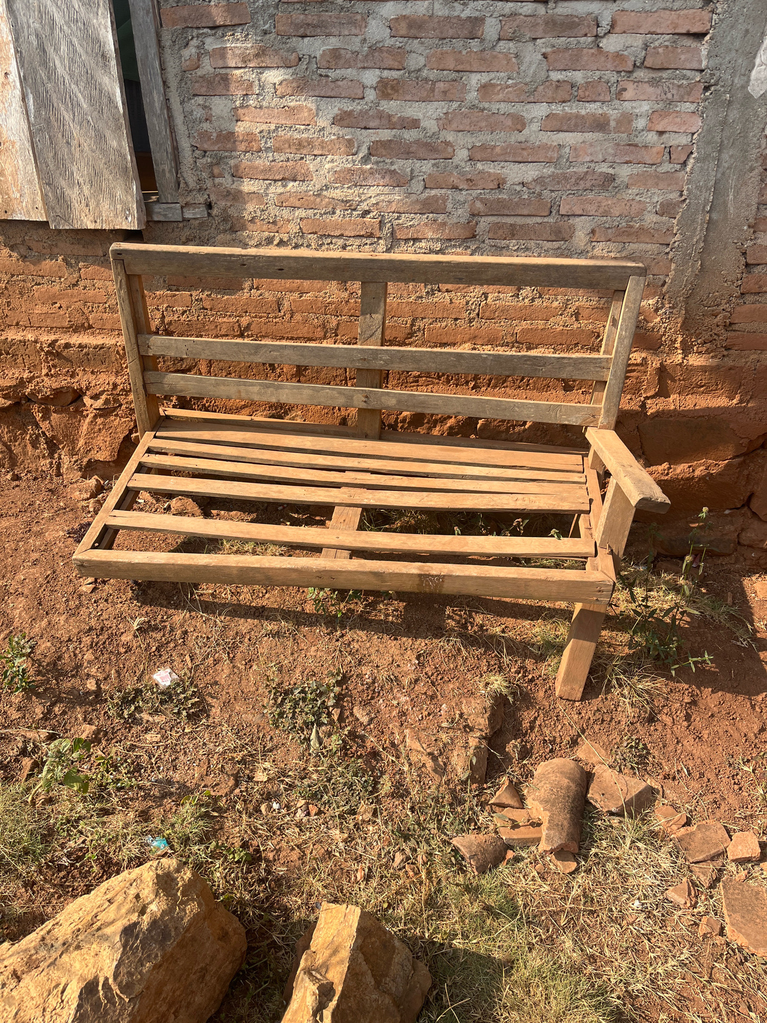 A rustic wooden bench is positioned in front of a weathered brick wall. The bench is constructed from rough-hewn planks and appears to be in a state of disrepair, with gaps between the slats of the seat. The brick wall behind it is made of red bricks with visible mortar, and part of a wooden window frame is visible on the left. The ground in front of the bench is reddish-brown soil with sparse dry grass and some scattered rocks. The scene appears to be outdoors, with natural sunlight casting shadows. There is no indication of human presence. The location is identified as Sidomulyo, Indonesia.