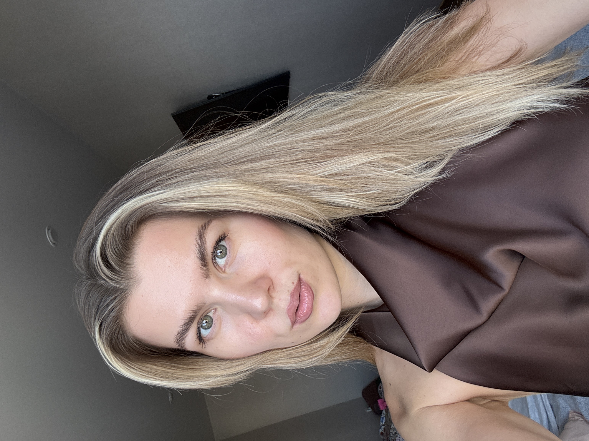 An outdoor, close-up, eye-level, medium shot shows a woman with blonde hair and green eyes. She is wearing a brown satin top. The background is a gray wall with a television mounted on it. The lighting is natural, coming from the left. The image is taken in an unknown urban location.