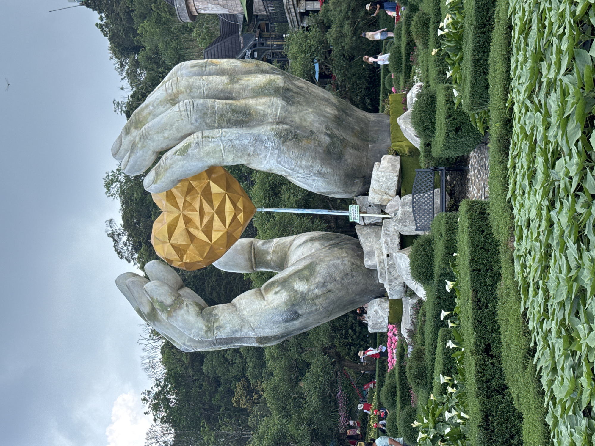 The image shows a sculpture of two large, aged-looking hands cradling a golden, geometrically patterned heart. The sculpture is situated within a garden setting with trimmed hedges and flowering plants. People can be seen walking in the garden. The scene appears to be from the Golden Bridge area of Ba Na Hills, Đà Nẵng, Vietnam, based on the garden design and surrounding landscape.