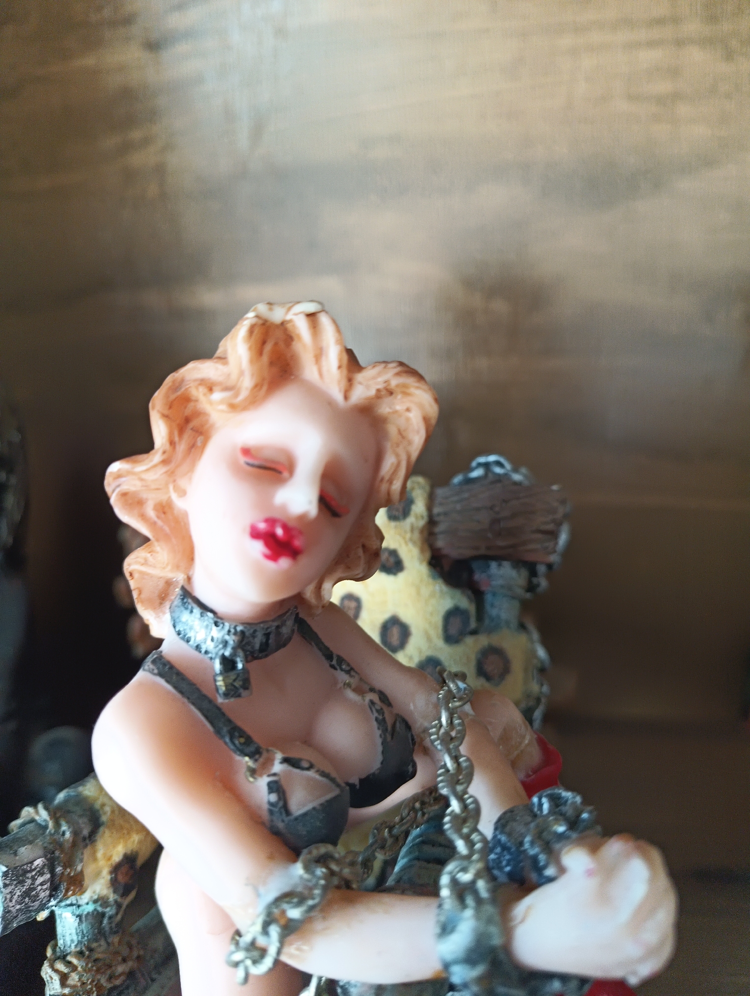 This is a close-up, eye-level, medium shot of a figurine of a woman. The figurine has fair skin, blonde wavy hair, closed red lips, and closed eyes. She is wearing a black bikini top, a black collar with a silver padlock, and several silver chains around her wrists and body. The background appears to be a wooden surface, possibly a shelf or wall. To the right of the figurine, there is a part of another object with a leopard print pattern and a dark, textured element. The lighting is soft, suggesting an indoor setting. There is no visible text in the image. The overall mood conveyed by the figurine's pose and accessories suggests a theme of captivity or restraint. The figurine is the primary subject, and the details of its attire and the chains are prominent. The setting is indistinct, focusing attention on the figurine itself.
