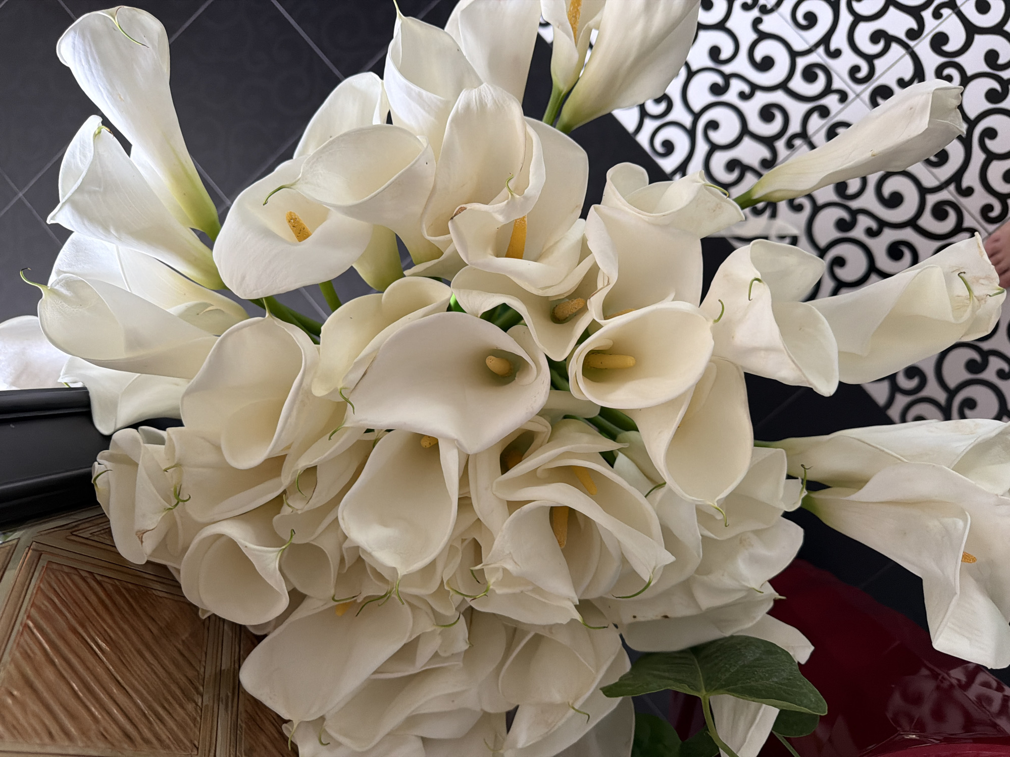 A bouquet of white calla lilies is arranged in a room, with a portion of a person's foot visible at the bottom right. The flowers are the primary subject, filling most of the frame. The background consists of patterned flooring and a dark surface. The overall impression is one of a decorative arrangement in an indoor setting.