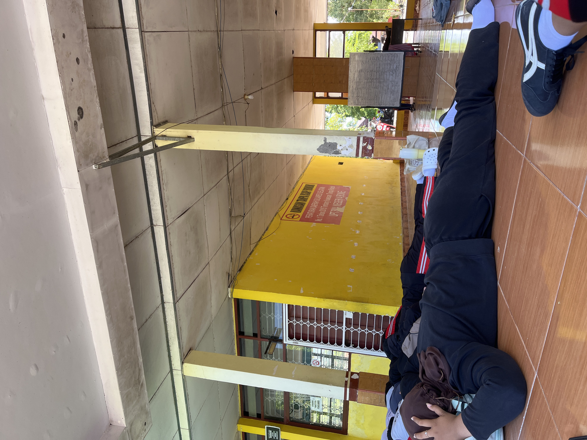 The image depicts a group of individuals lying on a tiled floor in what appears to be an outdoor or semi-outdoor covered area. The structure has a yellow-painted wall with windows and a red-striped door. A sign with Indonesian text is visible on the yellow wall, indicating "KAWASAN TANPA ASAP ROKOK" (No Smoking Area). The ceiling is made of panels and shows some signs of wear. The individuals are dressed in dark clothing, with some wearing headscarves. They appear to be resting or sleeping. In the background, a whiteboard with writing and some trees are visible. The scene is located in Pompanua, Indonesia.