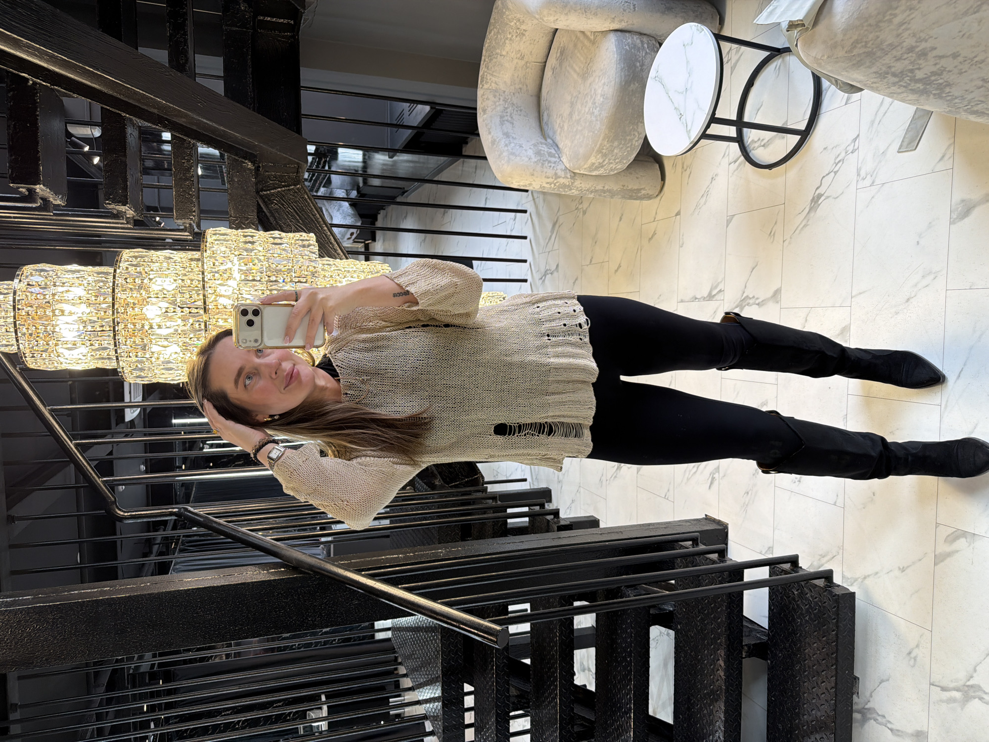 A woman is taking a selfie in what appears to be the lobby of a building. She is positioned near a black metal staircase, which is partially visible. Above her, a large, multi-tiered chandelier casts light. She is wearing a beige sweater with distressed detailing, black leggings, and black boots. Her right hand is raised to her hair, while her left hand holds a smartphone.
Behind her, marble flooring can be seen, along with furniture including a round white marble table and light gray armchairs.
It is not possible to determine the location in the city of Moscow, Russia, from this image alone.