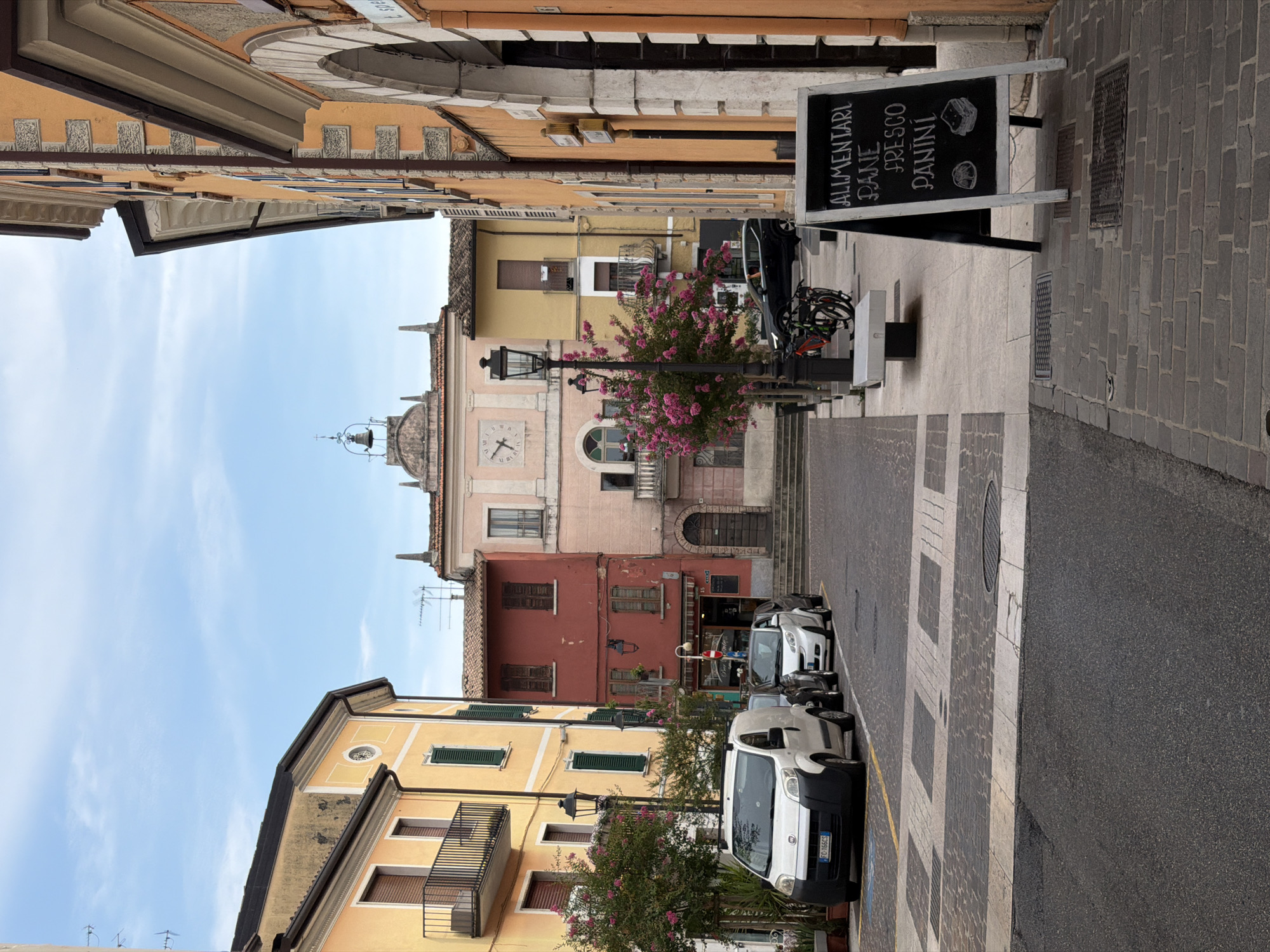 The image depicts a street scene in Padenghe sul Garda, Italy, on a cloudy day. The perspective is from a street level looking towards a plaza area. On the left, a yellow building with a balcony and dark shutters is visible. Adjacent to it is a red building with dark shutters and a street lamp. In the center, a larger pink building features a clock on its facade and a bell tower above. The street is paved with asphalt and a section of the plaza is made of cobblestones and decorative paving stones. Several cars are parked on the street, including a white van on the left and a white smart car and a dark SUV further down. A black A-frame chalkboard sign stands on the right sidewalk, displaying handwritten text that reads "ALIMENTARI PANE FRESCO PANINI" and drawings of bread and sandwiches. A black lamppost stands near the sign. In the background, bicycles are parked near a white bench. The buildings exhibit typical Italian architecture with arched windows and ornate details. The sky is overcast with some patches of blue visible. There are no people visible in the scene.