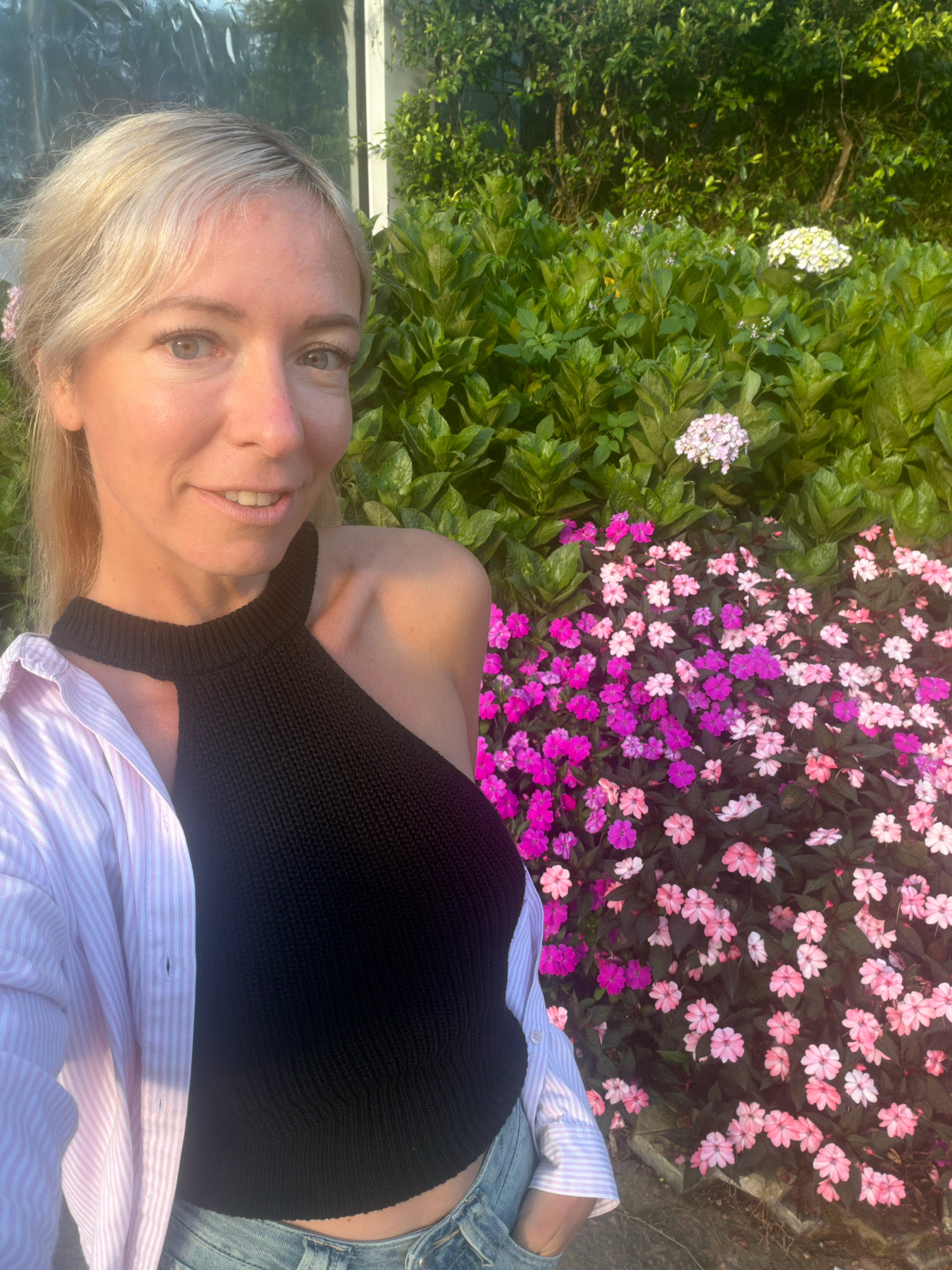 A woman with blonde hair and fair skin is taking a selfie in front of a dense green hedge with clusters of pink and white flowers. She is wearing a black halter-neck top, a light pink and white striped button-up shirt, and blue jeans. The lighting suggests it is daytime, possibly late afternoon, with the sun casting a warm glow. The background appears to be a garden or park setting. The location is identified as Xuan Truong Flower Garden, Da Lat, Vietnam.