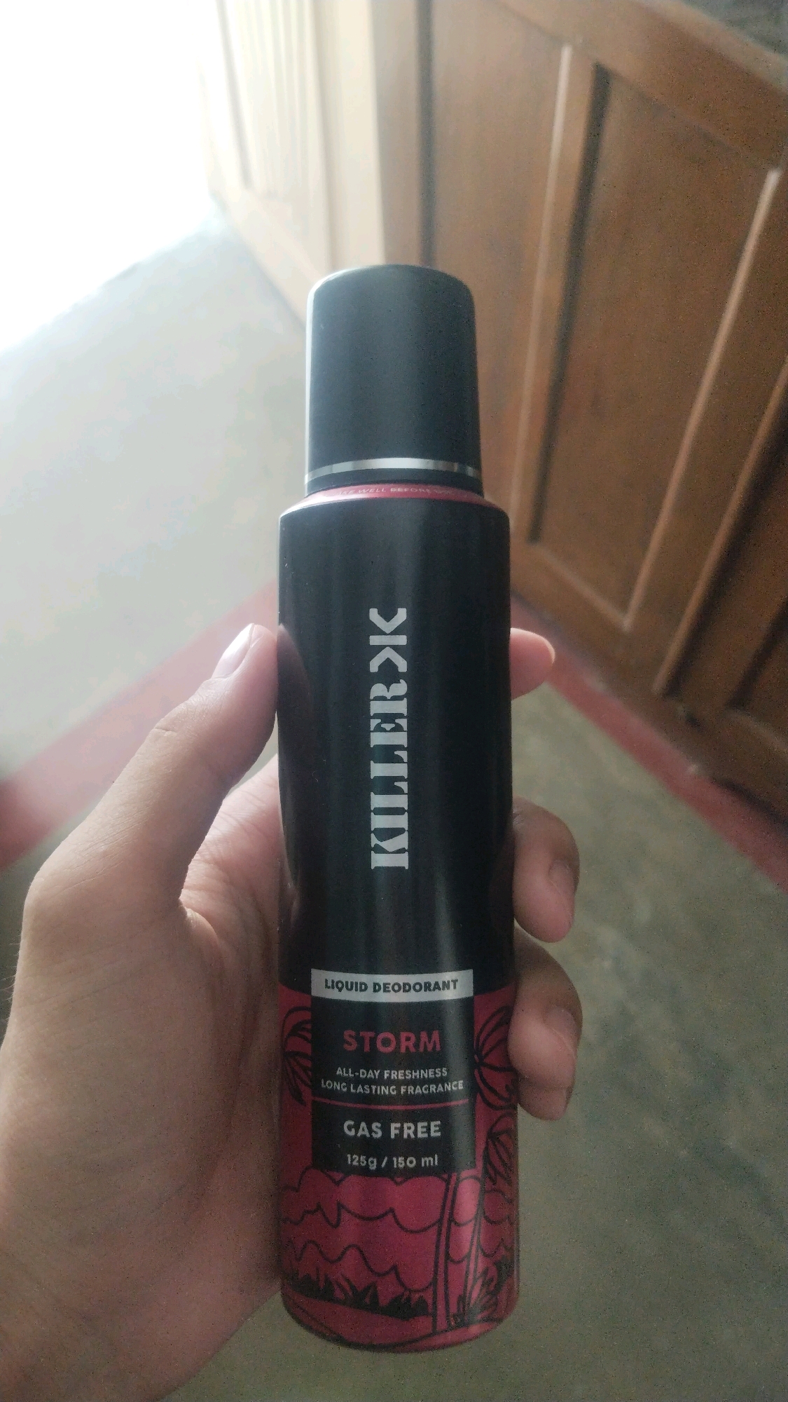 In Bihar Sharif, India, a person's hand holds a black aerosol can of Killer Storm Liquid Deodorant. The can features white text that reads "KILLERK" vertically along its side. The lower portion of the can displays "LIQUID DEODORANT" in white text on a black background, followed by "STORM" in large red letters. Below this, "ALL-DAY FRESHNESS" and "LONG LASTING FRAGRANCE" are written in smaller white text. The text "GAS FREE" appears in white, with "125g / 150 ml" below it. The can has a red and black patterned design near the bottom. The background shows a wooden door and a blurred floor, suggesting an indoor setting.