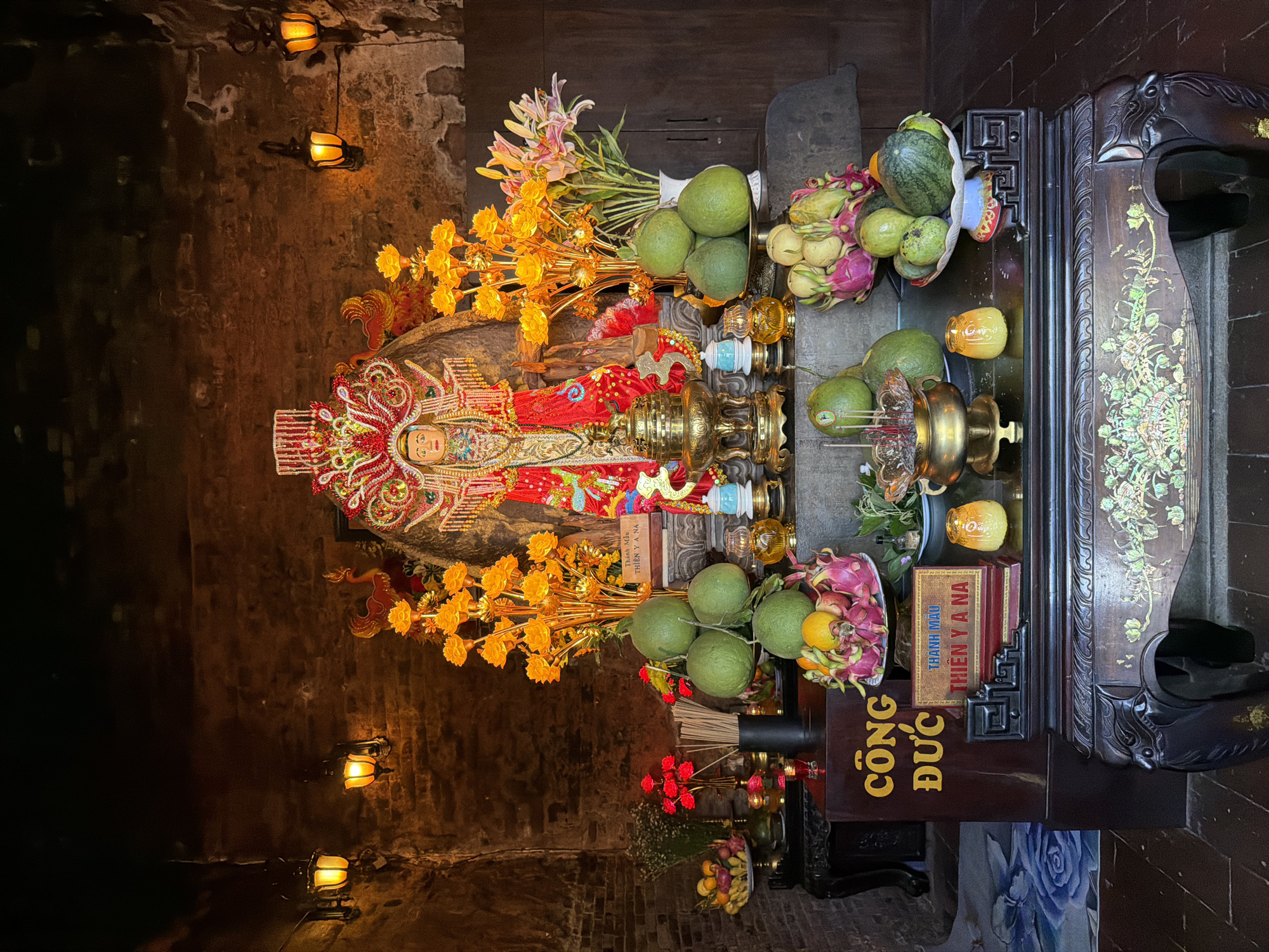 The image depicts an altar inside a dimly lit brick structure, likely a temple or shrine, located in Tuy Hoa Ward, Vietnam. The central focus is a richly adorned statue of a deity, possibly a goddess, wearing a red and gold robe and an elaborate headdress. Flanking the statue are tall, golden floral arrangements and a bouquet of pink and white lilies in a vase. In front of the statue, on a multi-tiered offering table, are various fruits including pomelos, dragon fruits, oranges, and a watermelon. Incense sticks in a brass urn and small candle holders with yellow flames are also present. To the left of the altar, a dark wooden cabinet displays more fruits and what appear to be red flowers in a vase. Two yellow lanterns hang on the brick walls, providing ambient light. A sign with Vietnamese text, "CÔNG ĐỨC THIÊN Y A NA," is visible on a dark wooden box in the foreground. The overall atmosphere is one of religious reverence and offering.