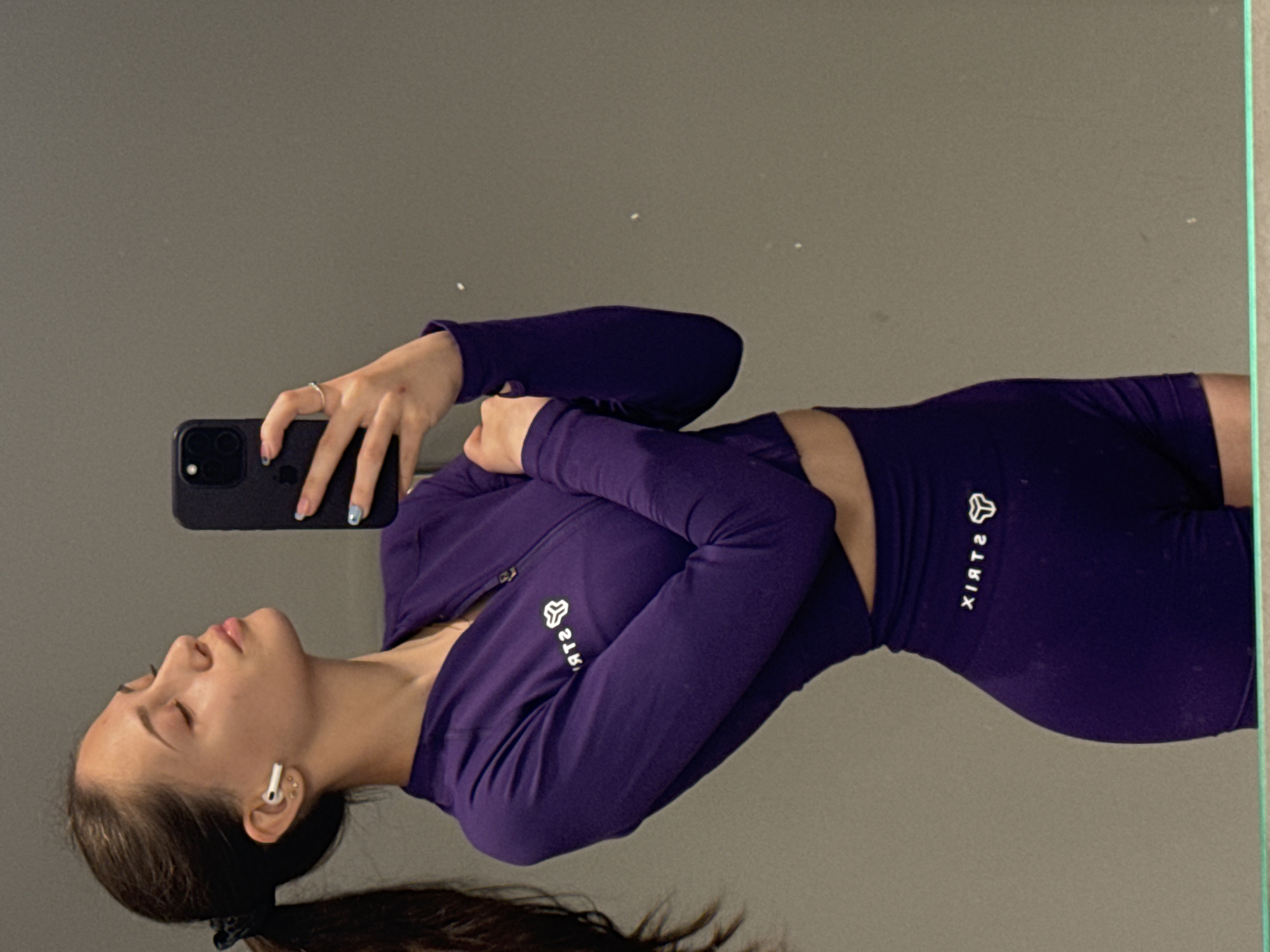 A young woman is taking a selfie in a mirror. She is wearing a matching purple workout set consisting of a long-sleeved zip-up jacket and shorts. The brand name "STRIX" is visible on both pieces. She is holding a black smartphone in her left hand and has an earpiece in her left ear. Her hair is pulled back into a ponytail. The background appears to be a plain, grey wall with the mirror reflecting the room. The exact location in Sofia, Bulgaria is not possible to determine from the image.