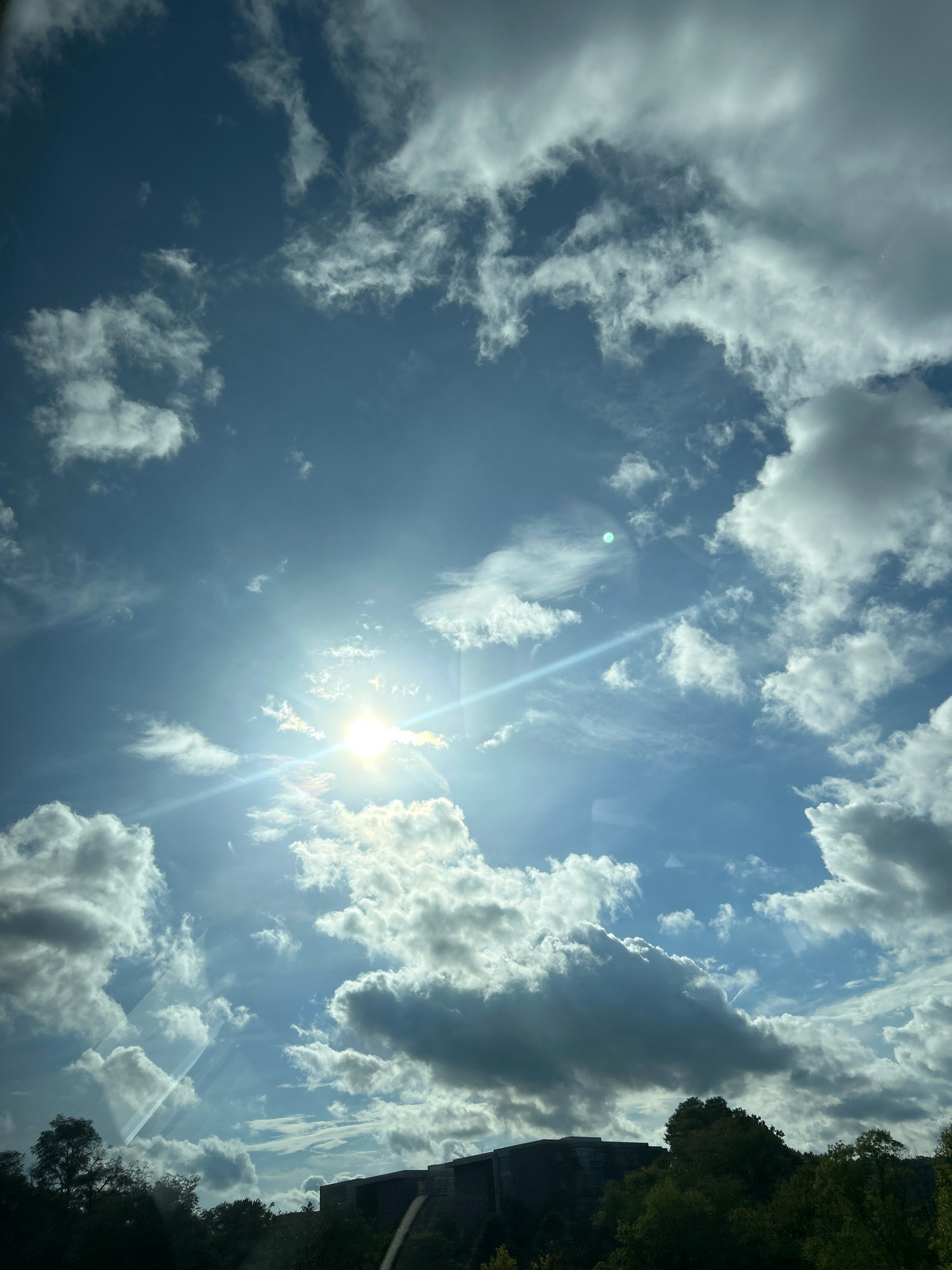 The image captures a sky filled with a mix of white, fluffy clouds and thinner, wispy ones against a bright blue sky. The sun is prominently visible, shining brightly and casting a lens flare across the frame, with a distinct blue glare to the left of the sun. Shafts of light are visible breaking through the clouds. In the lower portion of the image, the dark silhouettes of trees and the top of a building are visible against the bright sky. The building appears to have a modern, somewhat angular design. The scene suggests an outdoor setting, possibly during the daytime. The image's perspective is looking upwards towards the sky. The location is Collier Township, United States of America.