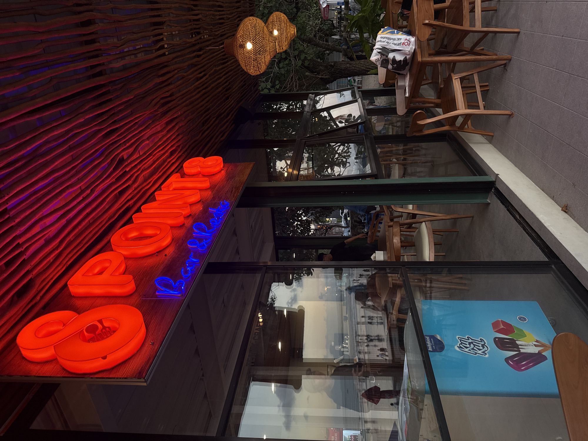 The image shows the exterior of a restaurant in Kuta, Indonesia, named "8 Points Bar & Restaurant". A large red neon sign with the restaurant's name is prominently displayed. The building has a modern design with large glass windows and a wooden slatted ceiling. Inside the restaurant, wooden tables and chairs are visible. In front of the restaurant, there is an outdoor seating area. A blue ice cream freezer with the brand "KUL KUL" is placed near the window. The background shows a beach scene with people walking along the shore and a body of water. Two woven pendant lights hang from the ceiling, illuminating the outdoor seating area.