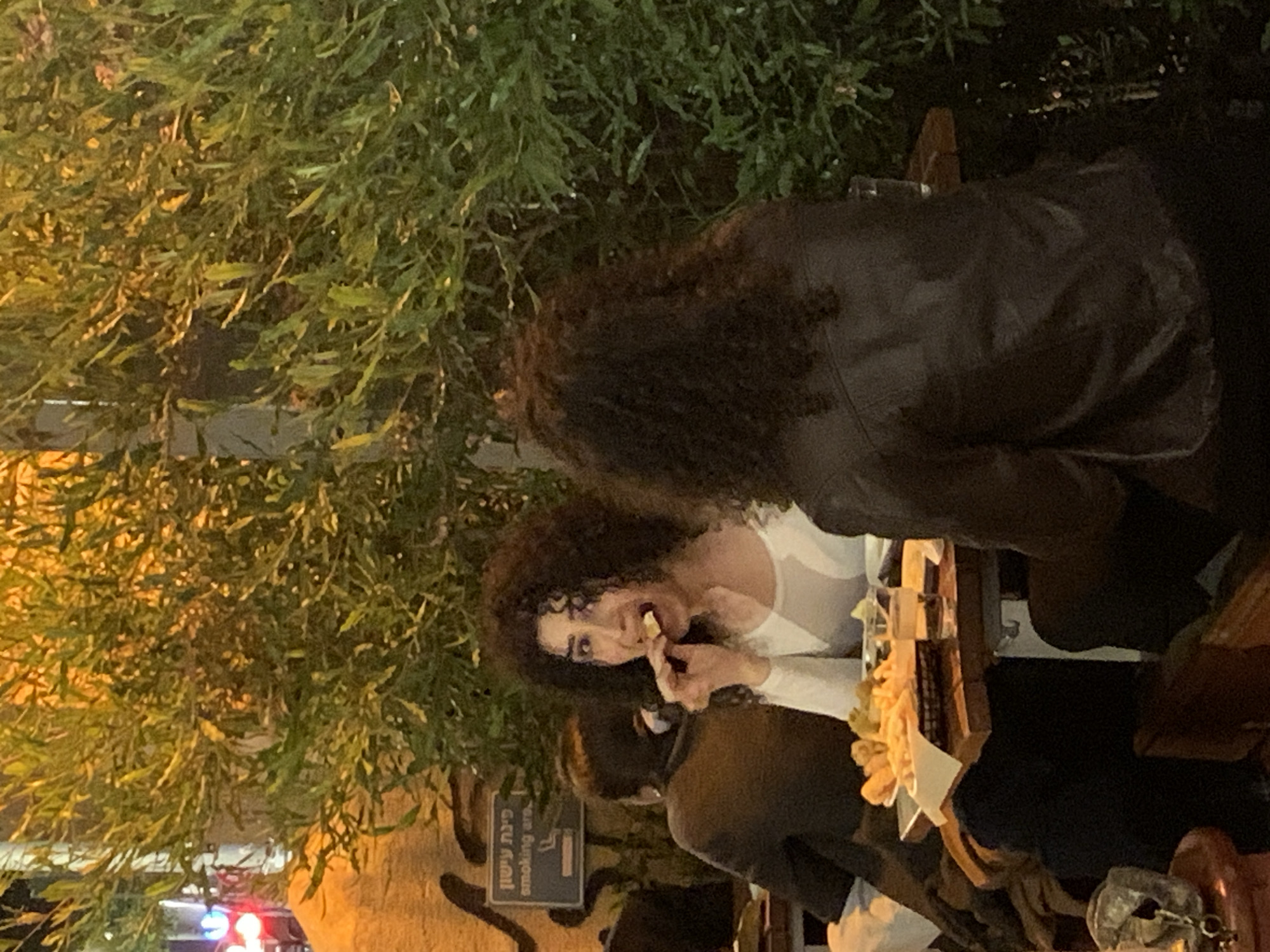 The image depicts an outdoor scene in what appears to be a public space, possibly a restaurant or café, in Tel Aviv, Israel. The focal point is a woman with curly hair, wearing a white top. She is in the act of eating something she holds in her fingers. Her attention is directed towards the food. She is seated at a table with other people. The scene includes two other people. One person is beside her, and another is in the background, wearing a dark leather jacket.
The background is filled with lush greenery, typical of Tel Aviv’s urban landscape, with a “Smoking area” sign. A decorative item is on the table.
The lighting suggests it is either evening or an overcast day. The presence of an outdoor seating area indicates a casual or social atmosphere.