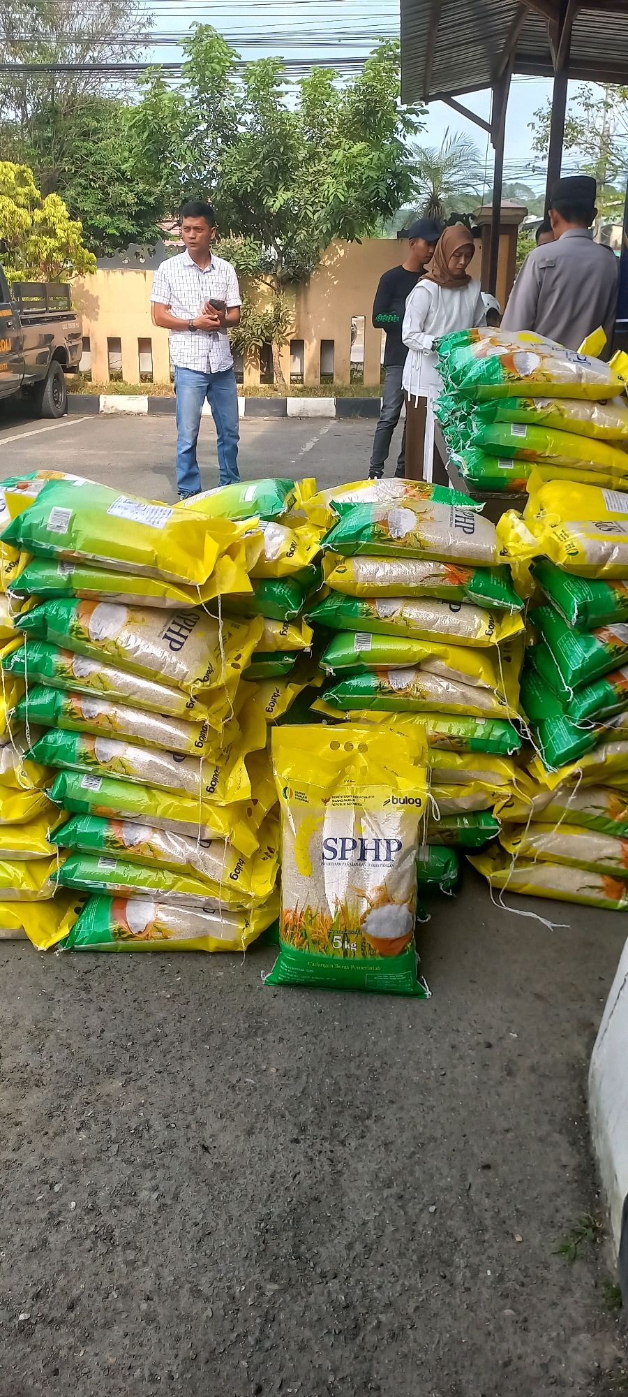 A medium shot captures a street-level view of a large stack of rice bags on an asphalt surface, likely in Samarinda, Indonesia. The bags, predominantly yellow and green, are piled high and appear to be a commercial product, with one prominent bag displaying the text "SPHP" and "5 kg," suggesting a government food program.
In the background, several individuals are visible. To the left, a man in a checkered shirt and jeans stands holding a smartphone. Behind him, a vehicle, possibly a truck with "POLISI" visible, is partially seen. Further back, a group of people, including a woman wearing a hijab and a white top, and another person in a black t-shirt, are gathered. To the right, a person in a light-colored uniform, possibly security or law enforcement, stands near an overhang structure.
The environment suggests an outdoor setting, with trees and vegetation visible in the mid-ground and a building structure in the background. The lighting indicates daylight. The overall scene depicts an organized distribution or collection of goods, with a focus on the stacked rice bags.