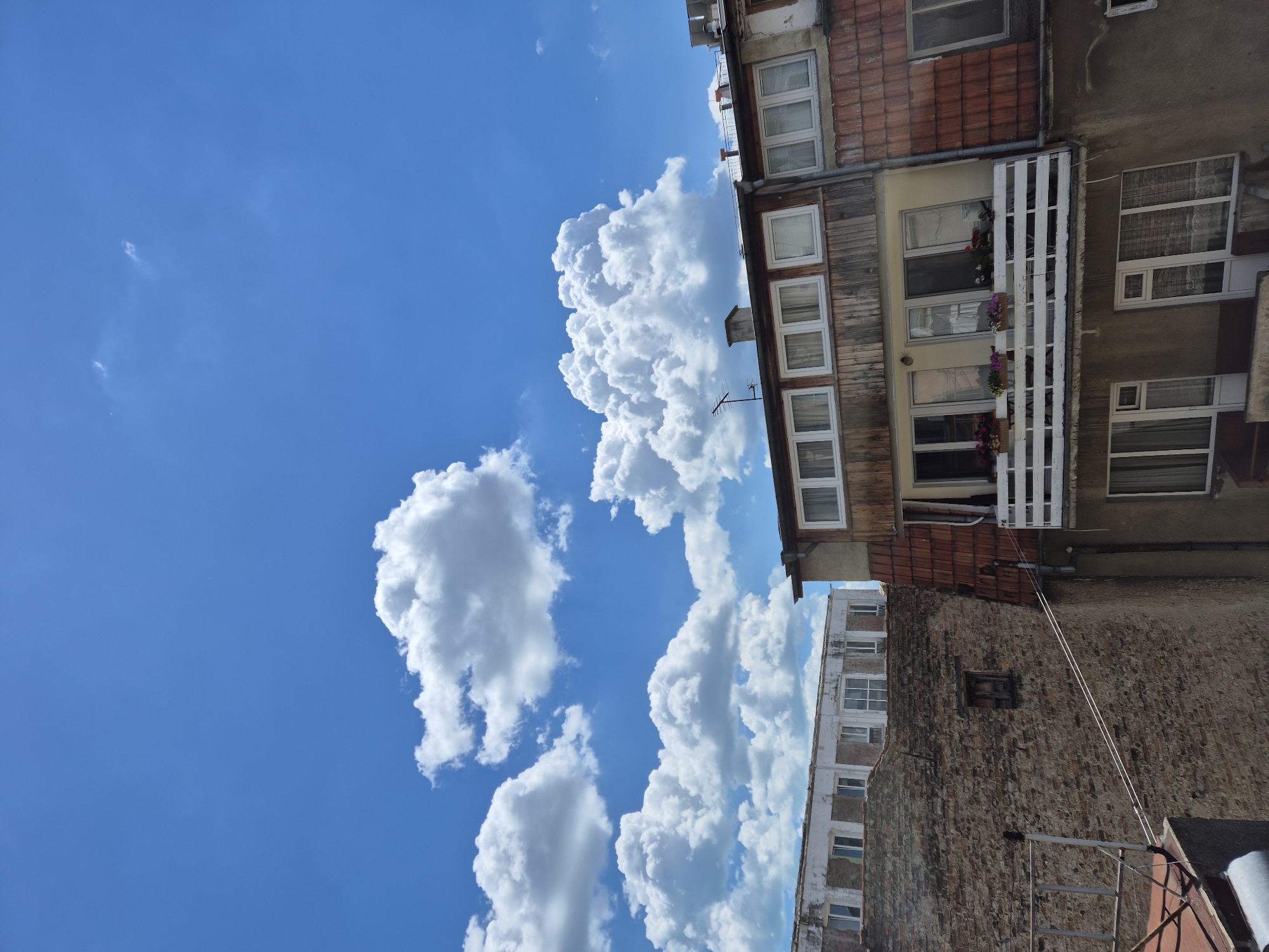 This is a photograph of buildings against a bright blue sky filled with fluffy white clouds, taken in Varna, Bulgaria. The perspective is from a high vantage point, looking down slightly at the rooftops and upper floors of several structures.
On the right, a multi-story building features a tiled roof and wooden paneling on its upper facade. A balcony with a white railing, adorned with flower boxes, juts out from one level. Below the balcony, more windows are visible. The building's lower section appears to be made of concrete.
To the left of this building is a section of a brick wall, appearing aged and textured. A small, dark, wooden window is set into this wall. Beyond the brick wall, another building is partially visible, with a lighter colored facade and multiple windows. The overall impression is an urban environment, possibly residential, with a mix of older and more modern construction. The sky dominates the upper portion of the image, suggesting a clear and sunny day.