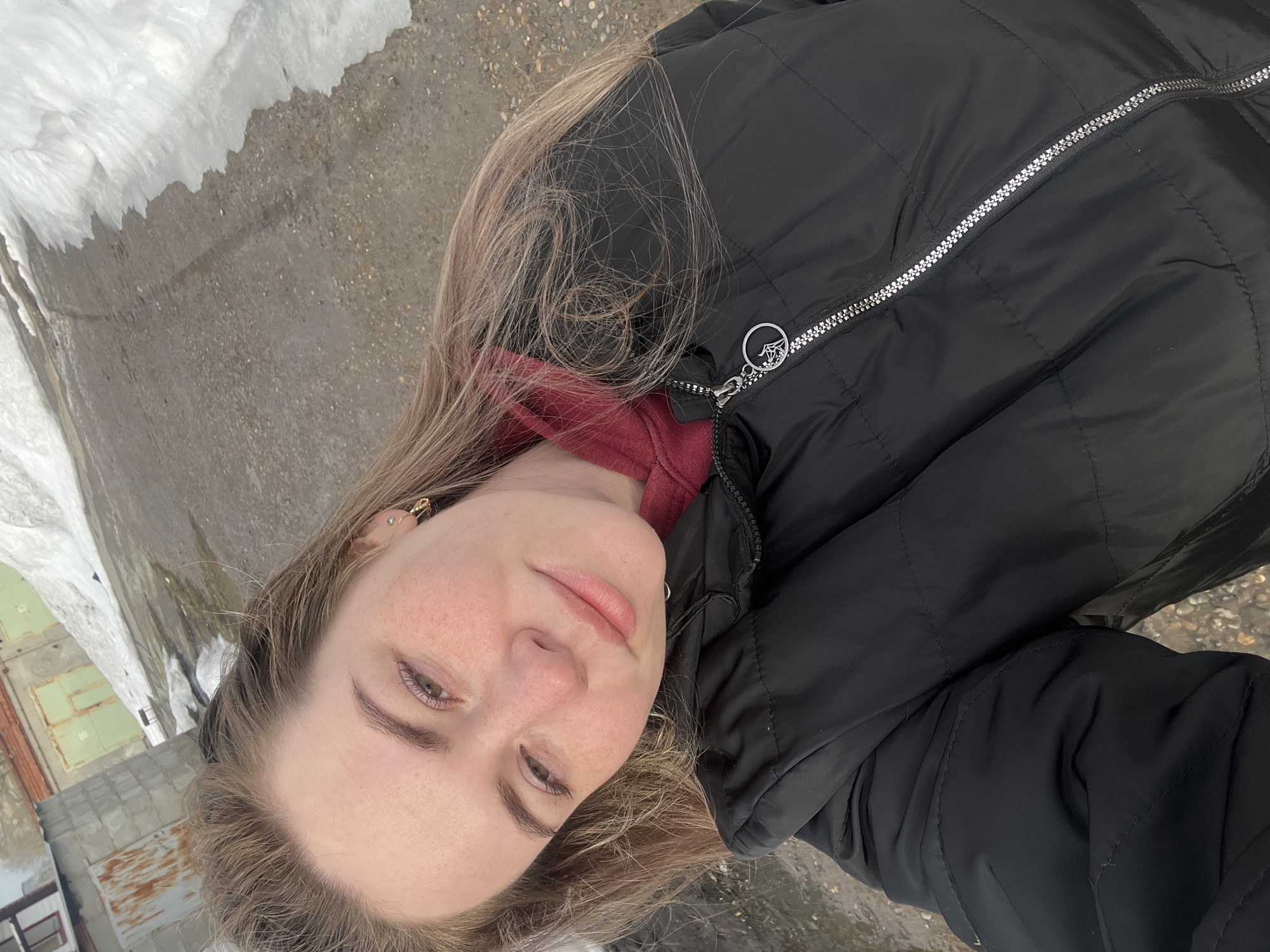 A young woman poses for a selfie outdoors. She wears a black puffer jacket over a red hoodie. Patches of snow are visible along a road or walkway. The location appears to be an urban or industrial area, possibly Chaikovsky, Russia, based on the background architecture, which includes a building with corrugated metal siding and small windows.