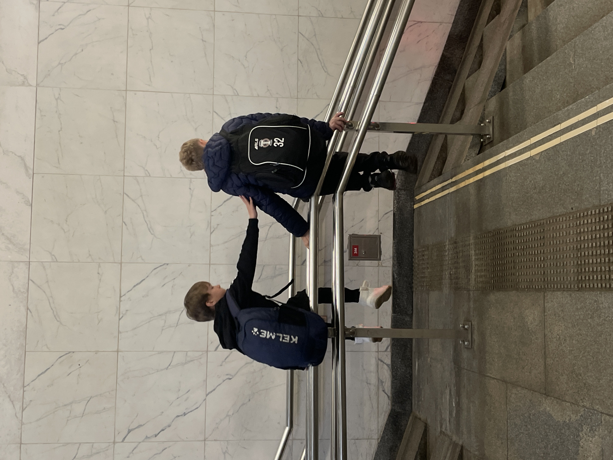 Two young boys are captured in an indoor setting, likely a public building such as a metro station or large hall, seen interacting on a stone staircase. The image is oriented vertically, with the boys positioned as if climbing or descending stairs.
The boy in the foreground, positioned higher on the stairs, wears a navy blue puffer jacket and a black backpack. The backpack features white text that reads "ARLS" above a circular logo and the number "32". He has light brown hair and is holding onto a silver metal handrail with his right hand. His left hand is lowered, seemingly in contact with the boy below him.
The second boy, positioned below the first on the staircase, wears a black hoodie and a blue backpack with "KELME" in white letters. He also has light brown hair and is extending his left arm upwards, possibly touching the first boy's arm or jacket. Both boys appear to be looking in each other's general direction, suggesting an interaction or shared activity, possibly playful, on the stairs.
The staircase itself is made of dark grey stone or concrete, with two thin, light-colored horizontal stripes running along the side wall. At the bottom of the visible stairs, there is tactile paving. To the left of the stairs, the floor is covered with large, light-colored marble tiles featuring subtle grey veining. A silver metal handrail with multiple parallel bars runs alongside the staircase. On the wall beside the stairs, a red utility box is visible, with the text "3WE" faintly discernible on it. The lighting is artificial, consistent with an indoor environment.