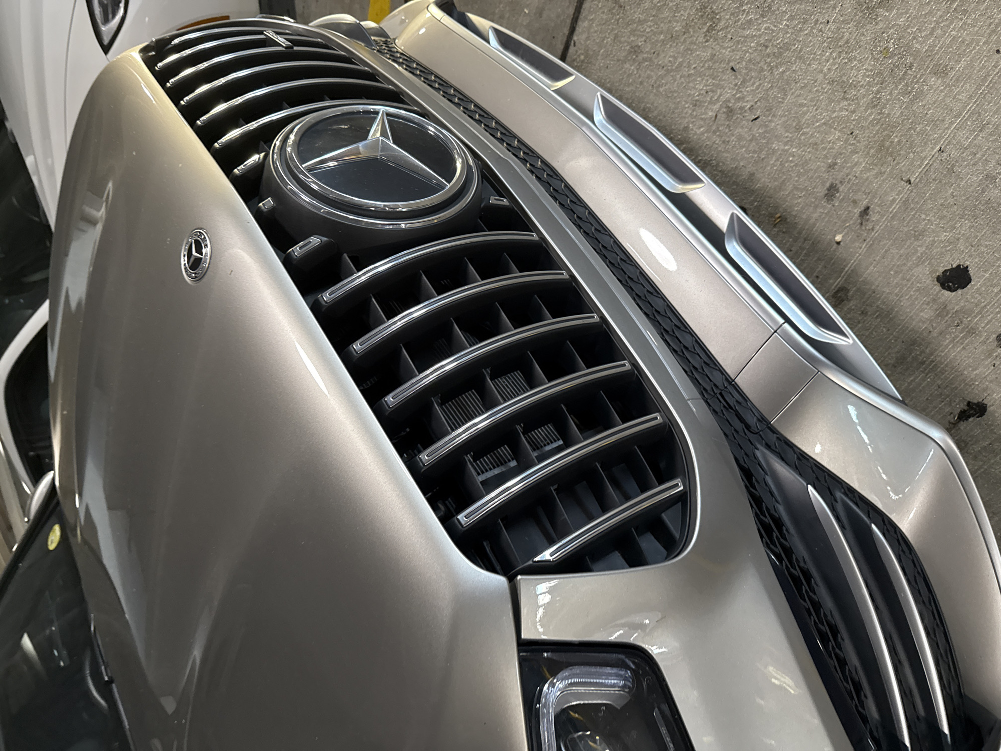 A close-up shot shows the front of a silver Mercedes-Benz, specifically the grill with the iconic Mercedes-Benz star emblem prominently displayed in the center. The grill features horizontal chrome bars. A smaller Mercedes-Benz emblem is also visible on the body of the car to the left of the grill. Part of the headlight assembly is visible at the bottom of the image. The car is parked in an enclosed area, possibly a garage or parking structure, with a concrete wall visible in the background. The location is Hollywood Seminole Reservation, United States of America.