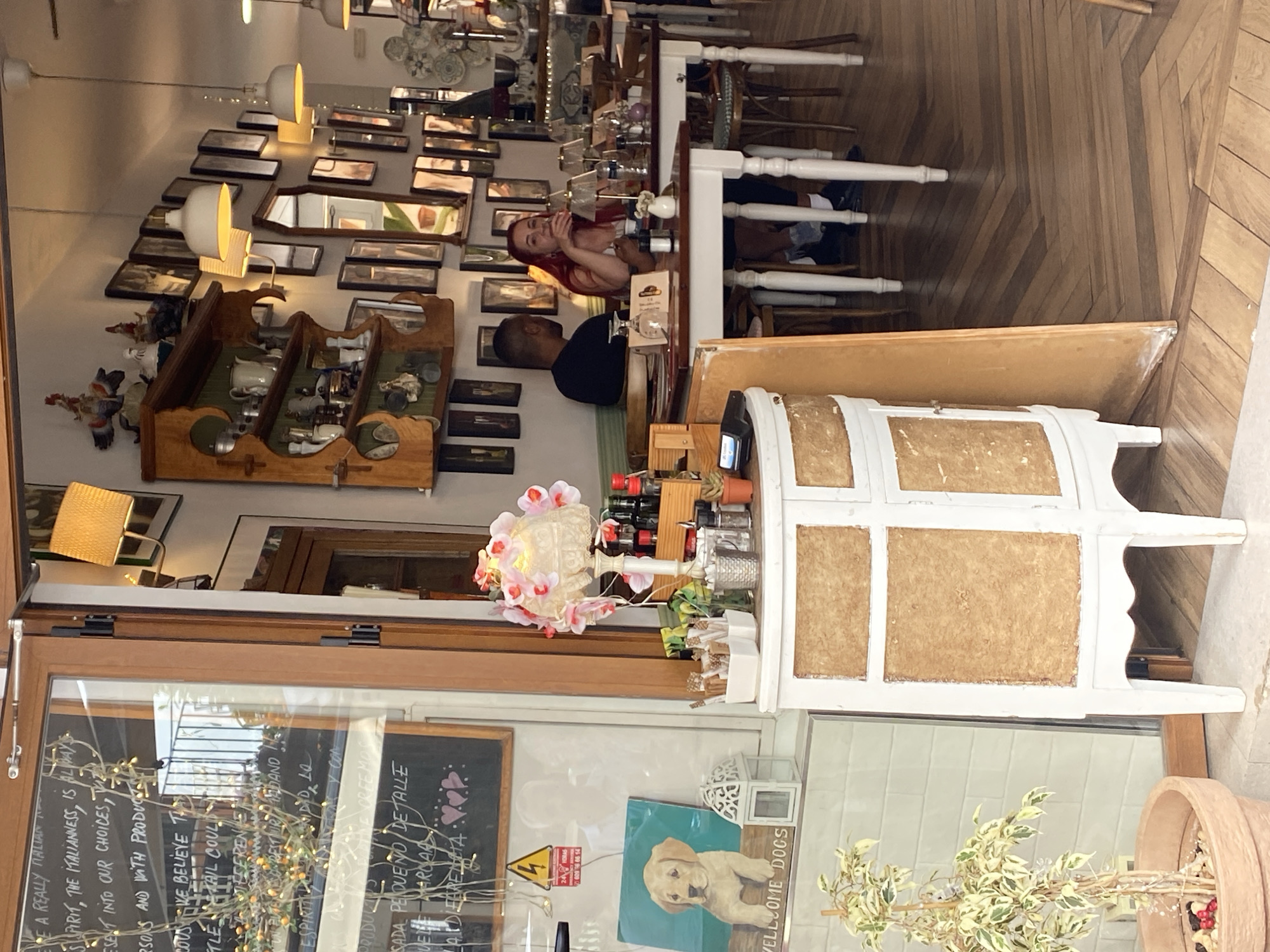 The image shows the interior of a restaurant in Nerja, Spain, seen from its entrance. Through the glass doors, a blackboard with handwritten text is visible, along with a painting of a dog and a decorative lantern. Inside, the restaurant features wooden flooring and tables with chairs. A couple is seated at one of the tables, with the woman visible from the waist up, talking while holding something to her mouth, and the man seated with his back to the camera. In the foreground, a white vintage cabinet with textured panels is placed near the entrance. Decorative lamps and shelves filled with various items are displayed on the wall. The lighting suggests it is daytime.