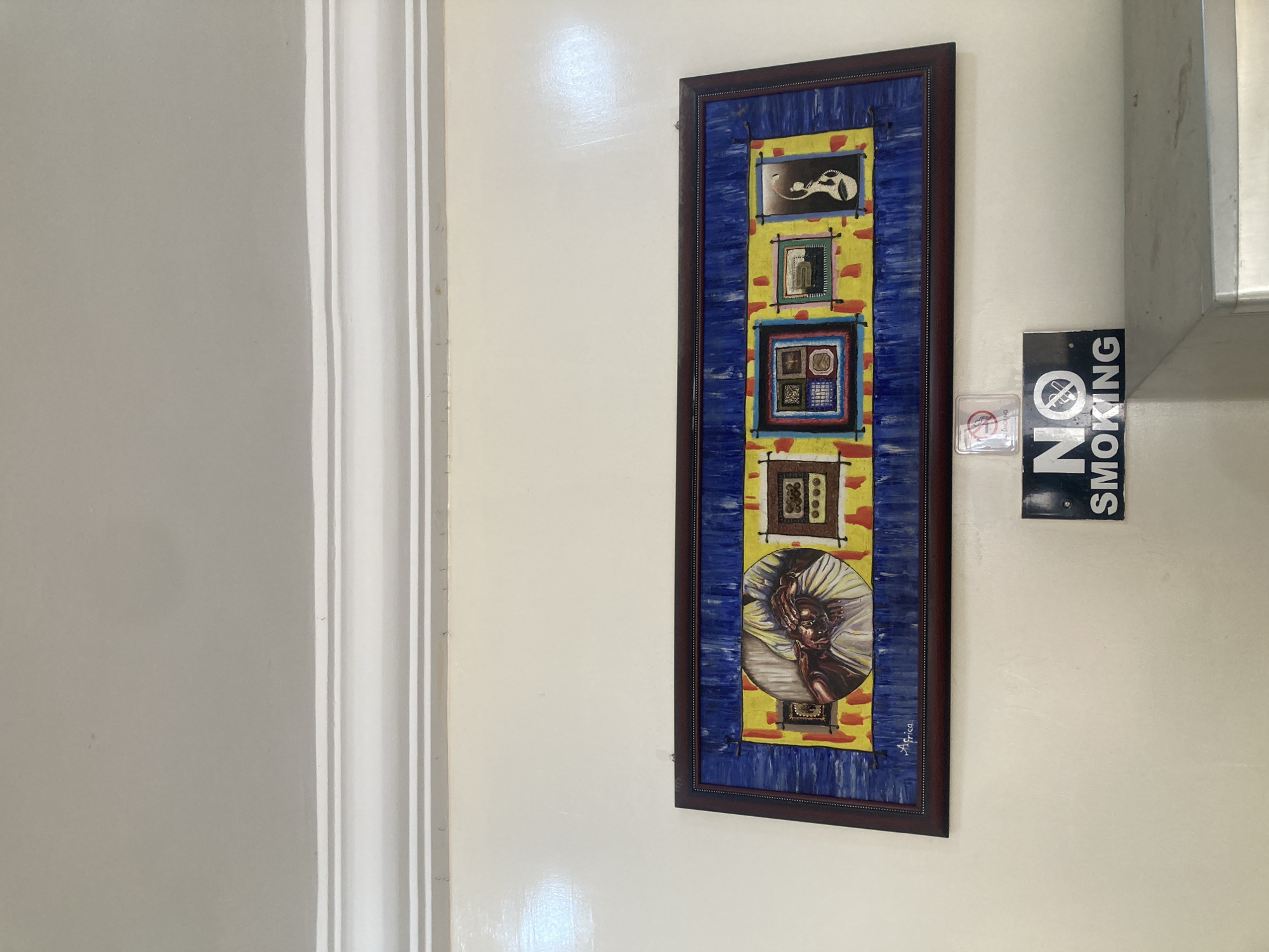 A framed painting hangs on a white wall, next to a "NO SMOKING" sign. The painting features a series of rectangular and circular vignettes with blue and yellow colors. The location is in Gura Top, Nigeria.