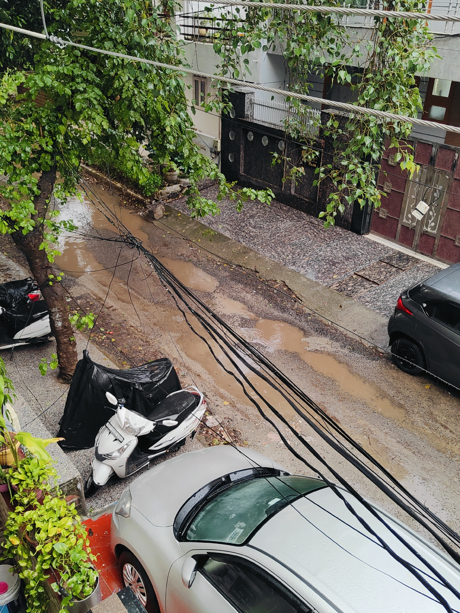 The image depicts a street scene in Gurgaon, India, during what appears to be a rainy day. The ground is wet and has several puddles. A silver car is parked in the foreground, its windshield showing raindrops. Next to it is a white scooter covered by a black tarp. Further down the street, another scooter is visible. There are also numerous black electrical wires crisscrossing the scene, some of which appear to be sagging. The surrounding buildings have architectural elements typical of urban India. Lush green trees with wet foliage hang over the street, indicating recent or ongoing rainfall. The overall impression is one of a typical monsoon day in an Indian city, with vehicles, infrastructure, and elements of nature coexisting.