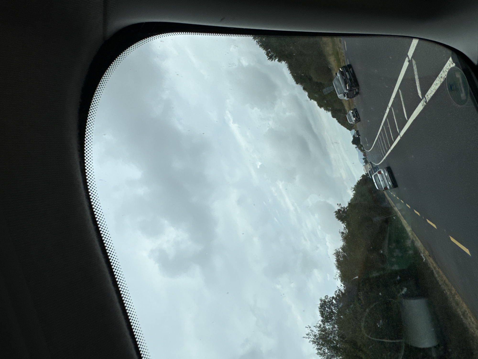 A grey, overcast sky is visible through a car windshield, looking out onto a multi-lane highway. Trees and vegetation line the right side of the road. Several cars are driving on the highway, including a dark SUV in the right lane and a silver sedan in the left lane further ahead. The highway has white lane markings and a yellow dashed line separating lanes. The image appears to have been taken while driving in or near Kilivory, Ireland, given the landscape and road features characteristic of the region.