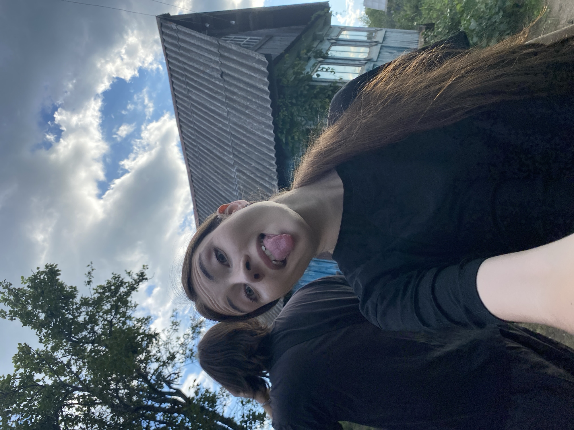 The image shows two people outdoors in what appears to be a rural setting, possibly Novyi Ural, Russia. A young person in the foreground is making a face at the camera, sticking their tongue out, with their head tilted sideways. They are wearing a black shirt. Behind them, another person, also wearing dark clothing, is visible from the shoulders up, with their head bowed.
In the background, an old, weathered wooden house with multiple windows is partially visible. The roof of the house is made of corrugated metal. Green foliage from trees and bushes surrounds the house and the people. The sky is cloudy with patches of blue, suggesting it might be daytime with mixed weather. The overall impression is casual and candid, likely a selfie or a photo taken by a friend. There is no visible text in the image.