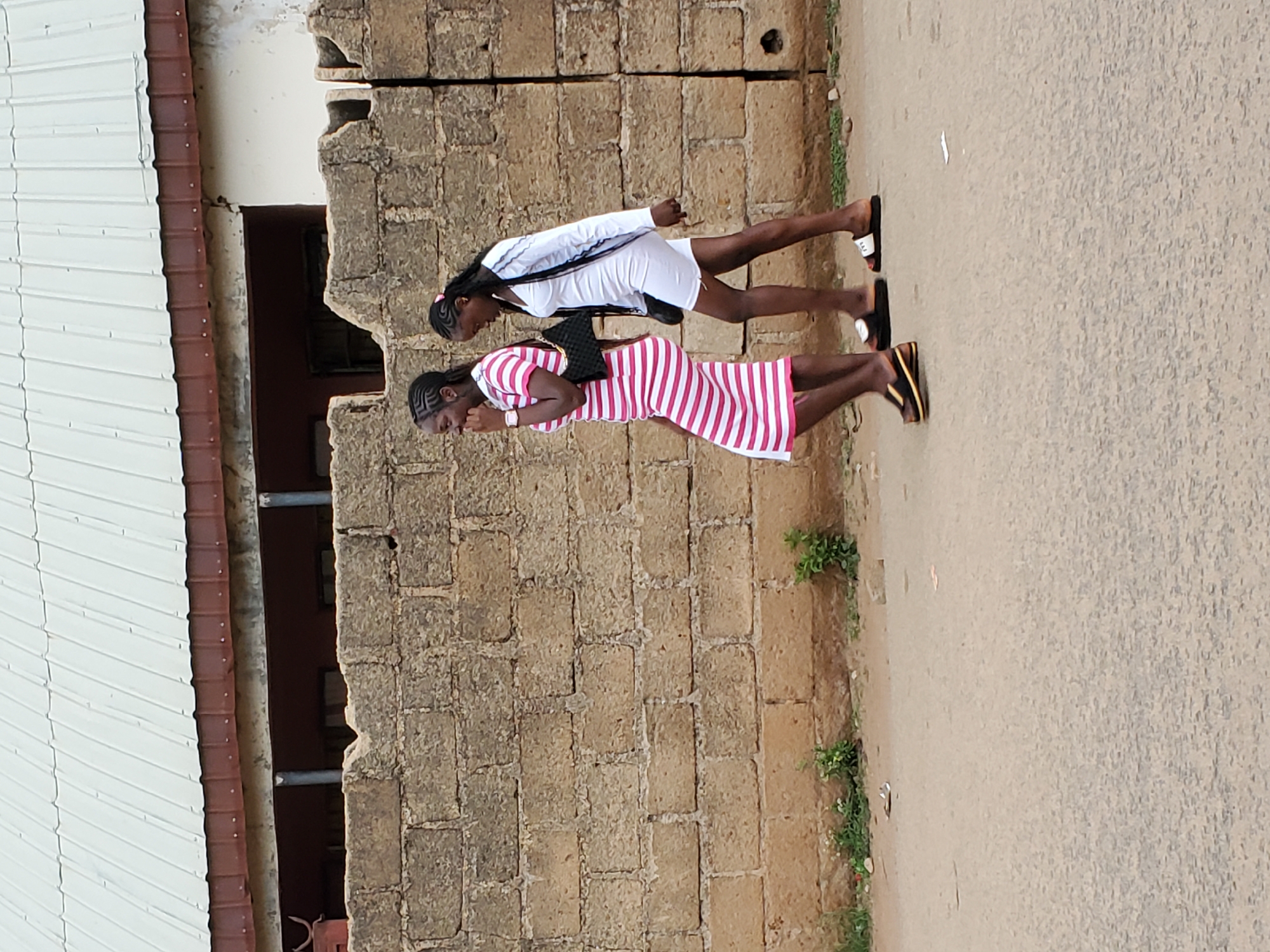 Two young women walk together along a street in Jos, Nigeria. The woman in the foreground wears a red and white striped dress, while the other is in a white dress and jacket. They are walking next to a stone wall and building. The road is paved and worn. The weather appears to be daytime.