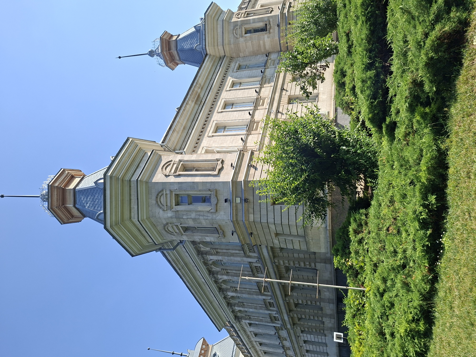 This is a medium-shot, low-angle photograph of the exterior of a light-colored, stone building with ornate architectural details. The building features multiple turrets with conical roofs, decorative cornices, and numerous windows. In the foreground, there is a lush green lawn and dense shrubbery. The sky is clear and blue. The building is the Palace of the Republic, located in Chișinău, Moldova.