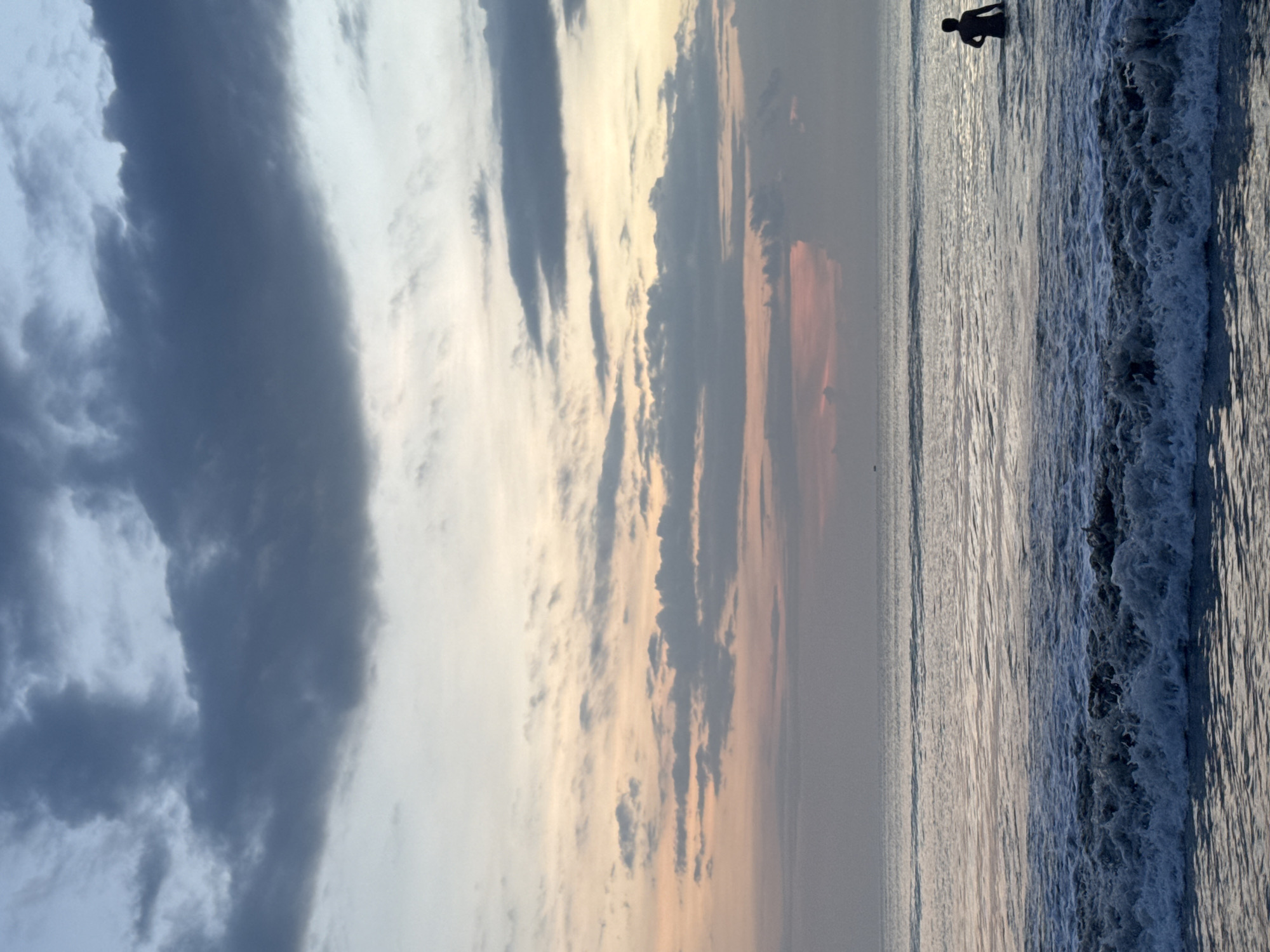 The image depicts a beach scene at sunset, presumably in Kuta Selatan, Indonesia. The sky is filled with various cloud formations, ranging from dark grey cumulus to light, wispy cirrus clouds tinged with orange and pink hues, indicating the setting sun. Below the sky is the ocean, with a person silhouetted against the horizon standing in the water. A wave is breaking on the shore to the right of the frame, with its foamy crest clearly visible. The water surface reflects the colors of the sky. The photograph is rotated 90 degrees counterclockwise.
