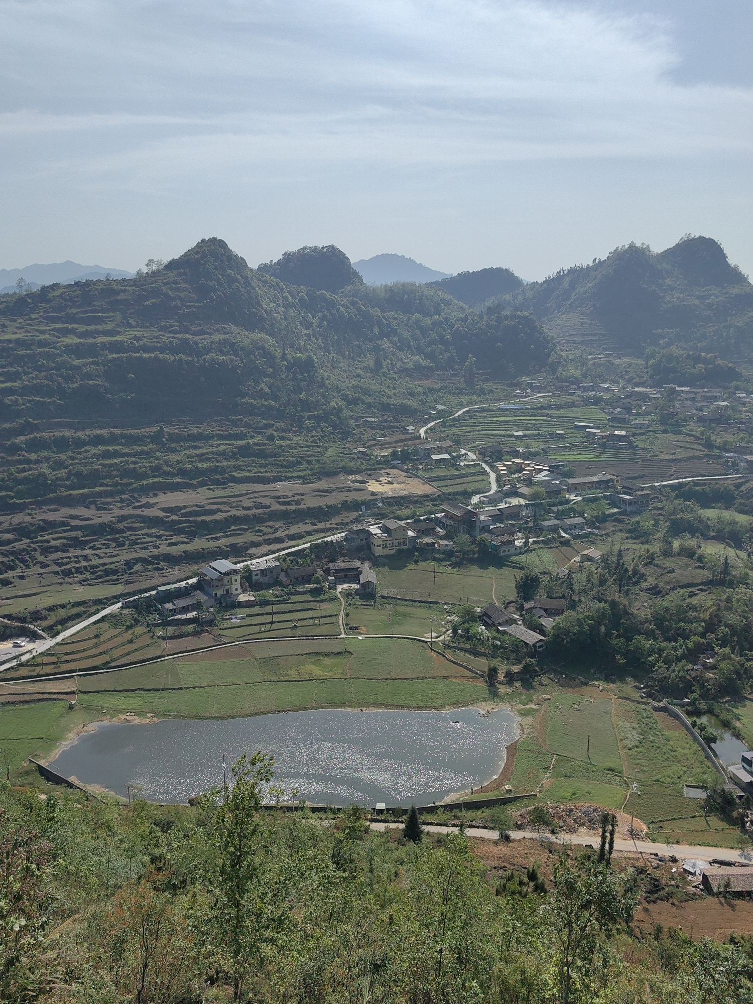 The image shows a panoramic view of the Lũng Cú Commune in Vietnam. A body of water, possibly a lake or reservoir, sits in the foreground, reflecting sunlight. Patches of cultivated land, appearing as green and brown fields, surround the water. A road runs along the side of the water, connecting some of the homes. A cluster of dwellings, appear to be homes, are visible further up the hillside. The hills in the background are terraced and covered in vegetation. A winding road connects the houses and runs up into the mountains. The sky is overcast.