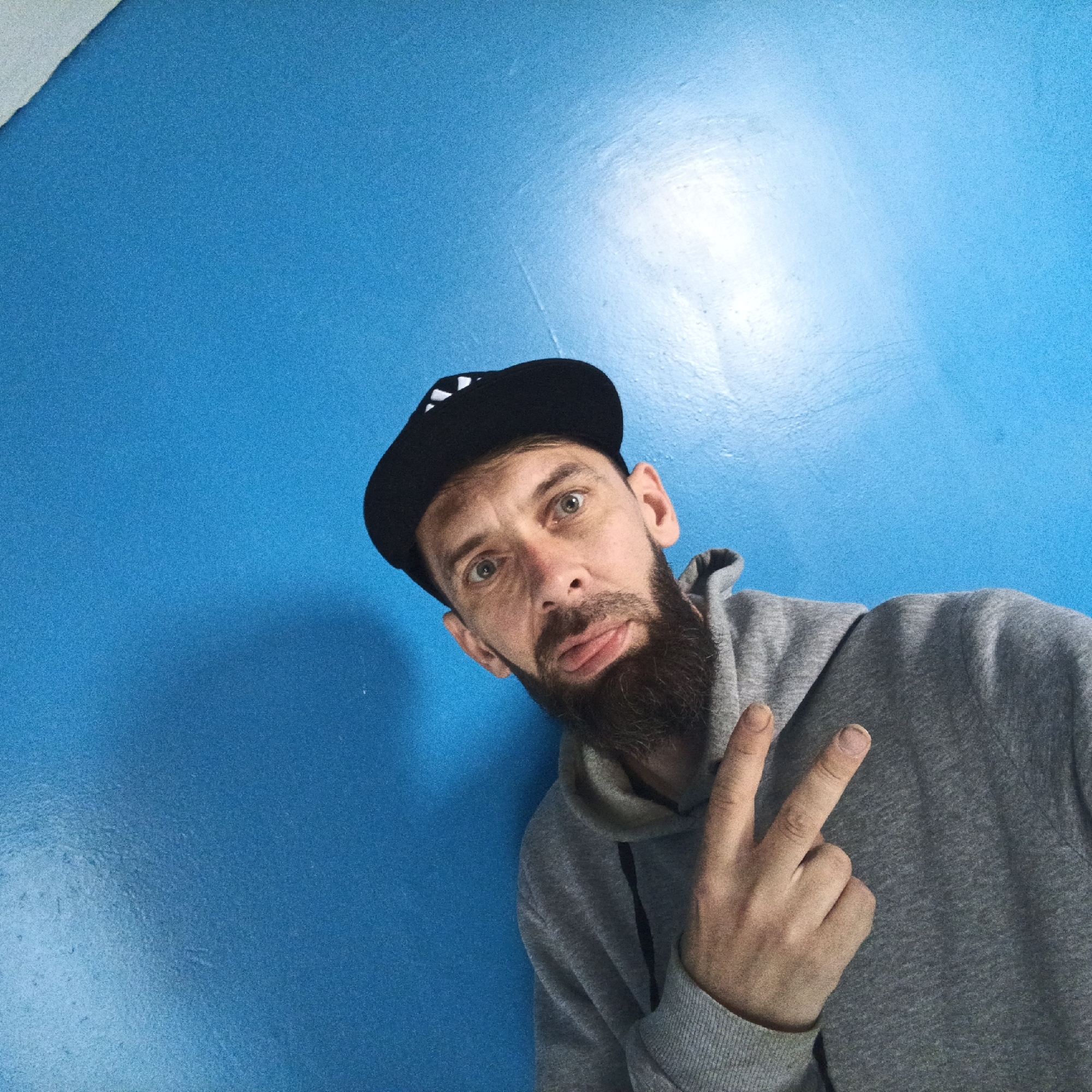 A man with a beard and a black baseball cap is shown in a selfie taken against a bright blue wall. He is wearing a grey hooded sweatshirt. The man is holding up two fingers in a peace sign with his right hand. The image appears to be taken indoors in an urban environment. There is no indication of the specific location being Марковский, Russia.