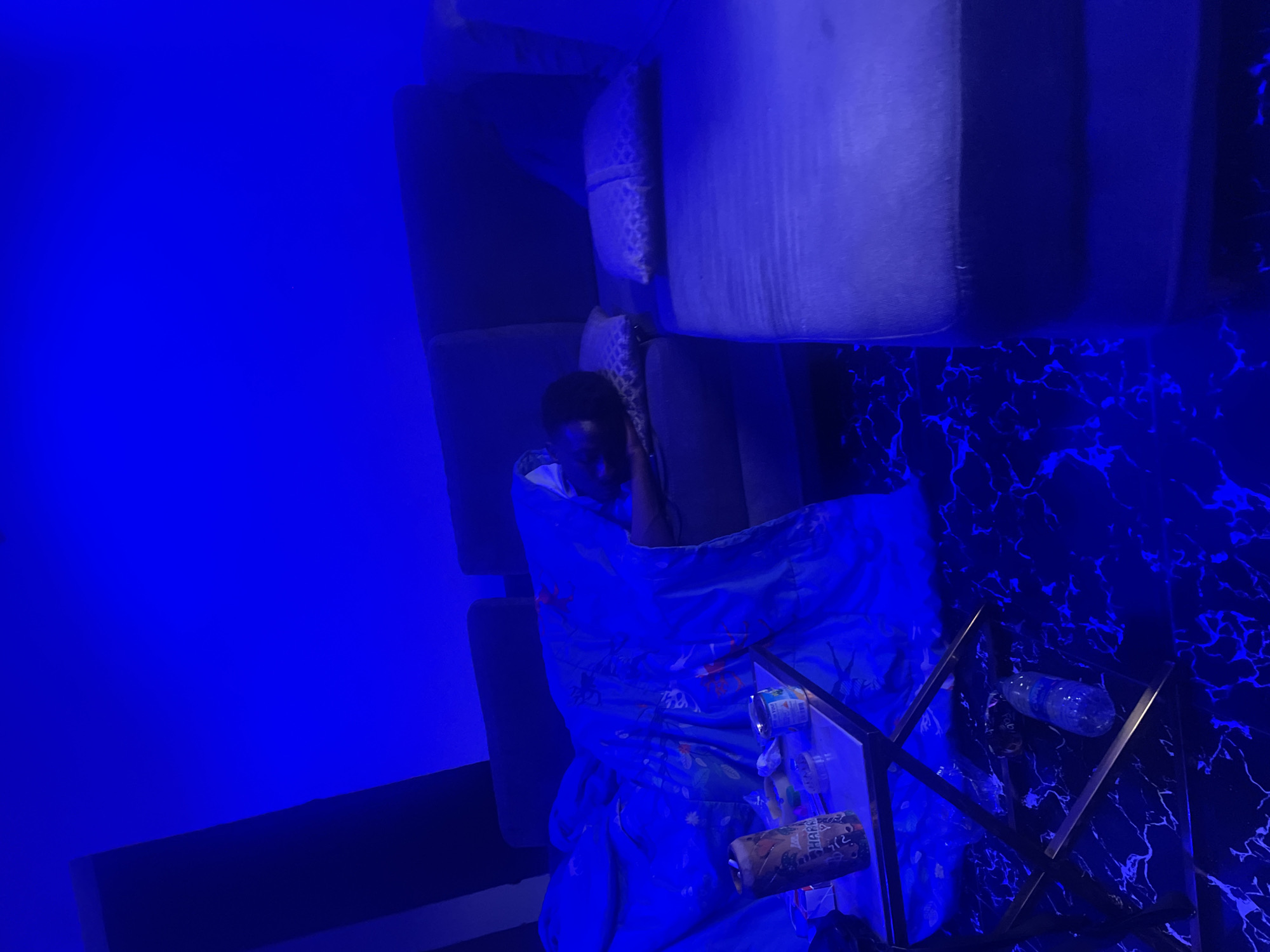 The image captures an indoor scene bathed in vibrant blue ambient lighting, likely signifying evening or nighttime. A young male is depicted resting or reclining on a dark-colored sofa, partially covered by a blue blanket adorned with a subtle pattern. His head and upper body are visible, and he appears to be looking towards the left of the frame.
To the right of the person, a side table with a distinctive black surface featuring striking blue or white marble-like veining is visible. The table's base consists of a metallic, geometric frame. Resting on this table are several items: multiple beverage cans or bottles, one of which shows partially discernible text that appears to be "HARRA" (possibly part of a brand name), and a clear plastic water bottle lying on its side. The surrounding walls are uniformly illuminated by the blue light, contributing to a serene and atmospheric setting. The scene suggests a moment of relaxation or repose in a living room or lounge, possibly in Abuja, Nigeria.