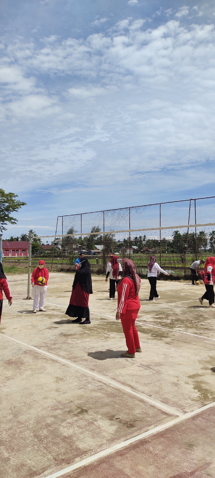 In Peurupok, Indonesia, a group of women are engaged in a volleyball game on an outdoor court. The court is made of concrete with white boundary lines. A volleyball net is stretched across the middle of the court. Several women are visible in athletic attire, mostly in red and white. One woman in the foreground, wearing a red tracksuit and headscarf, is looking towards the net. Another woman in a black abaya and headscarf stands near the center of the court, holding a volleyball. Further back, other women are positioned on the court, some appearing to be playing, while others observe. In the background, there is a fence, some trees, and buildings, suggesting an area adjacent to a residential or community space. The sky above is bright blue with scattered white clouds, indicating fair weather.