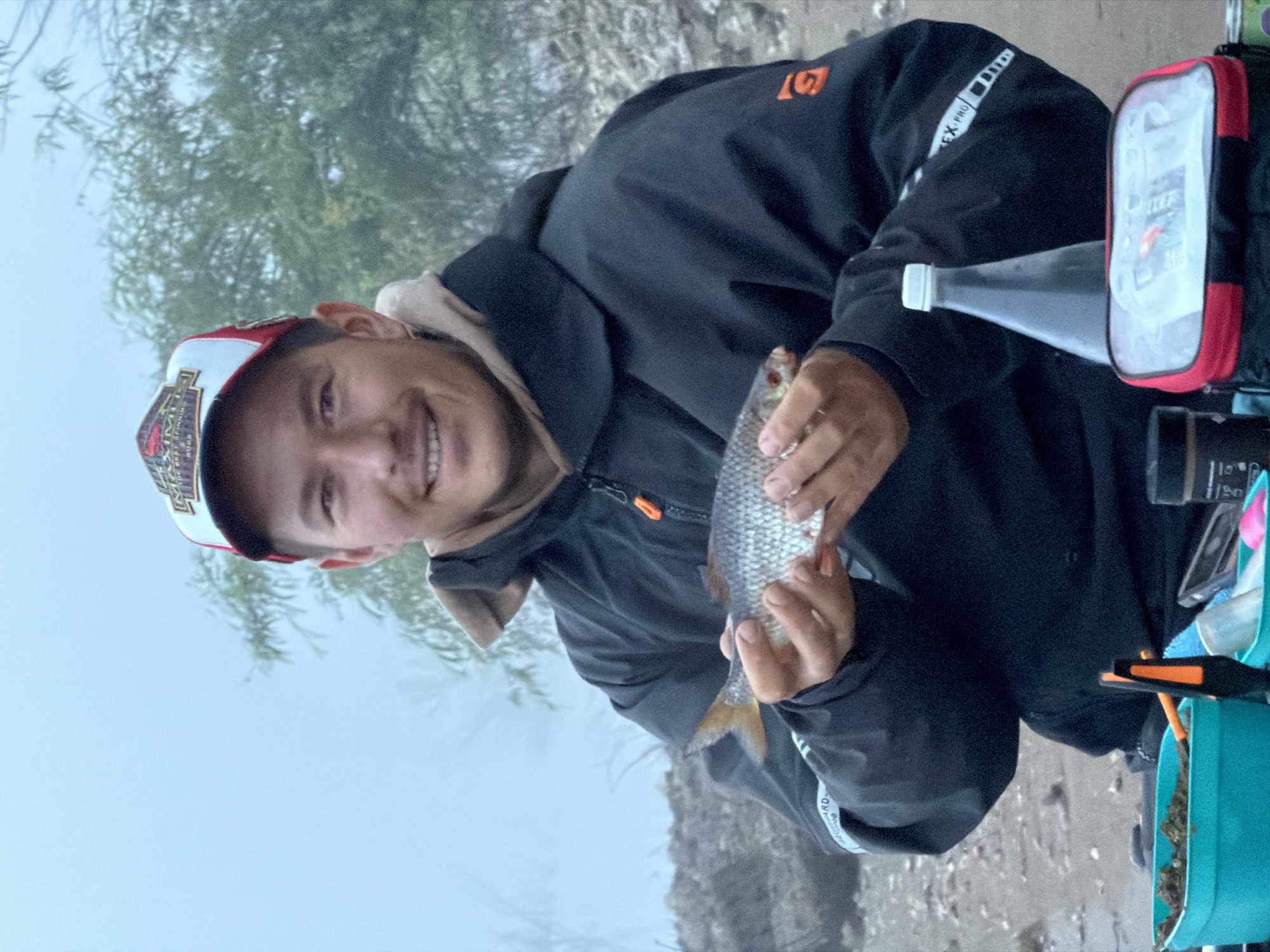 An outdoor, mid-shot image captures a young man, likely in Chaikovsky, Russia, joyfully holding a small fish. He is smiling broadly at the camera, his face conveying satisfaction. He wears a dark, possibly waterproof jacket with an orange zipper pull and a light-colored hooded sweatshirt underneath. A white and red baseball cap is on his head, featuring "MAXIM" and other unreadable text on the front.
The man holds a small, silver-scaled fish with both hands, its tail fin exhibiting a yellowish hue. To his right, several fishing items are visible: a clear plastic bottle with a white cap, a red and clear zippered pouch or bag with the word "ICE" discernible through the plastic, and a turquoise bucket or container filled with green material, likely bait.
The background is softly focused, revealing a natural landscape. A body of water, possibly a river or lake, is directly behind him, reflecting the overcast sky. Further back, green foliage from trees or bushes adds to the serene outdoor setting. The ground nearby appears to be rocky or muddy.
The lighting is soft and diffused, characteristic of an overcast day or early morning/late afternoon, with no harsh shadows. Visible text on his jacket sleeve includes "EX-PRO" and a stylized "G" or similar logo on the chest. The scene depicts a successful and pleasant fishing outing.